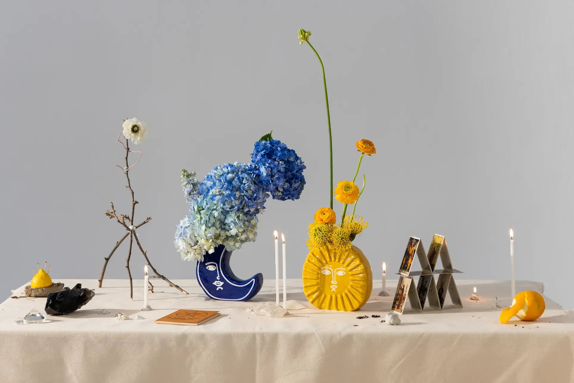 A decorated table features blue hydrangeas in a crescent moon vase, yellow flowers in the DOIY Sun Vase by DOIY, candles, crystals, a house of cards, and small objects on a white cloth—ideal for creative home decor.