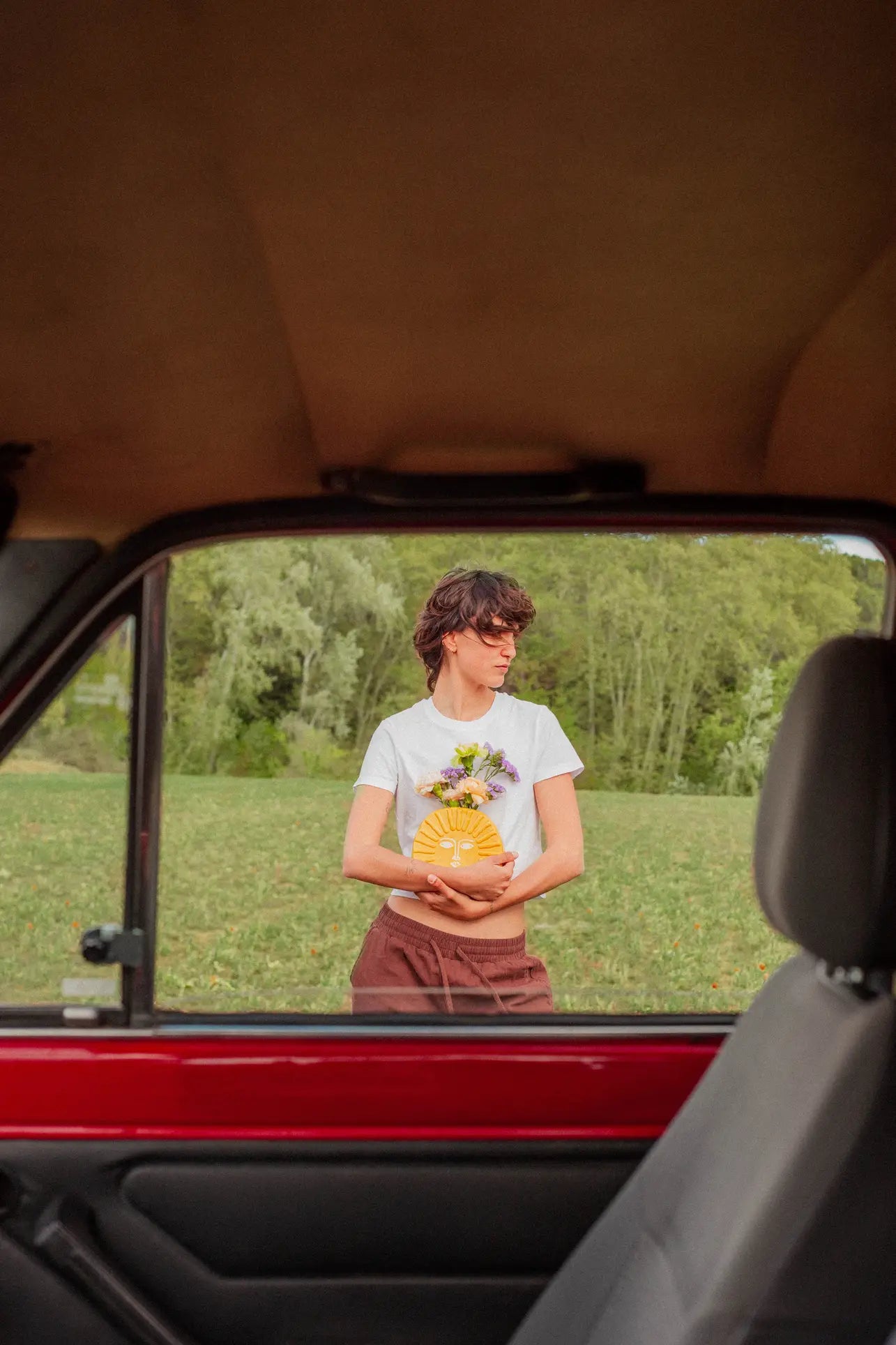 A person in a white T-shirt and brown pants stands by a red car, holding flowers ideal for the DOIY Sun Vase by DOIY. Theyre outdoors, with grass and trees visible through the open window.