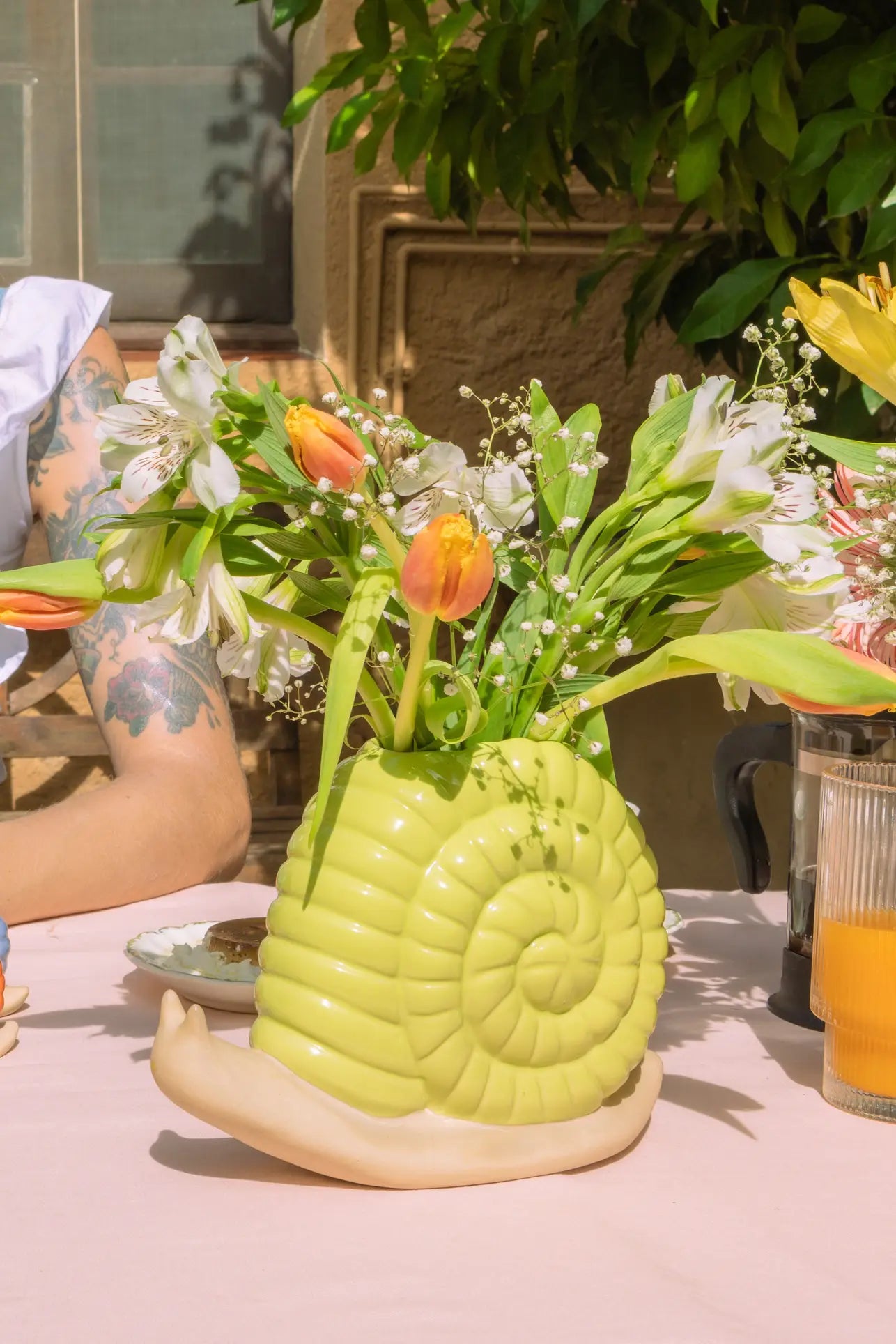 The DOIY Woodland Snail Vase by DOIY, featuring a yellow snail silhouette, holds orange tulips and white flowers on an outdoor table beside a tattooed arm and a glass of orange juice, with green leaves and a window in the background.