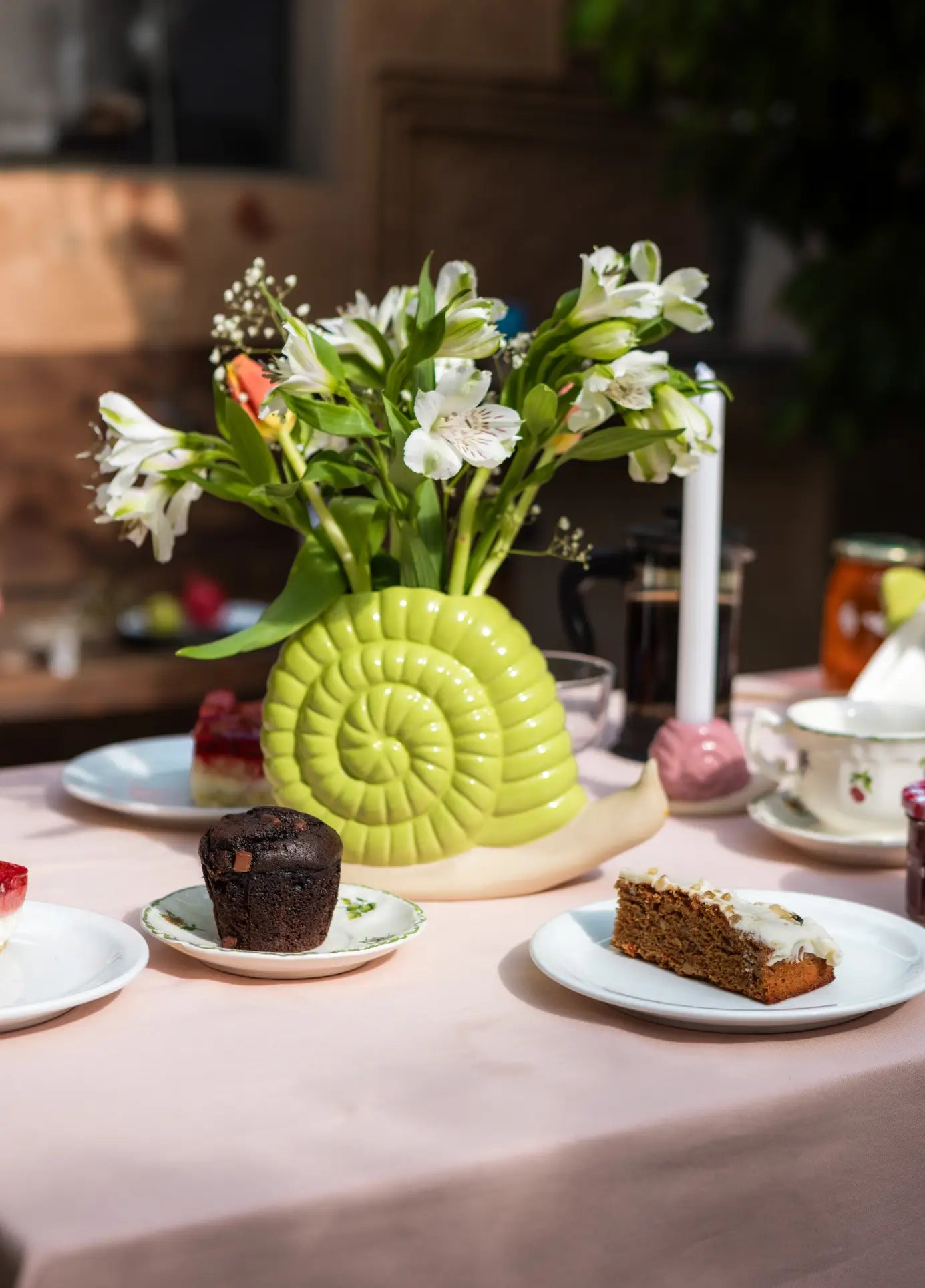 A table bathed in sunlight features the DOIY Woodland Snail Vase by DOIY, its yellow ceramic form holding white flowers—a charming accent among cakes, a chocolate muffin, lit candle, and teacups on a pink tablecloth.