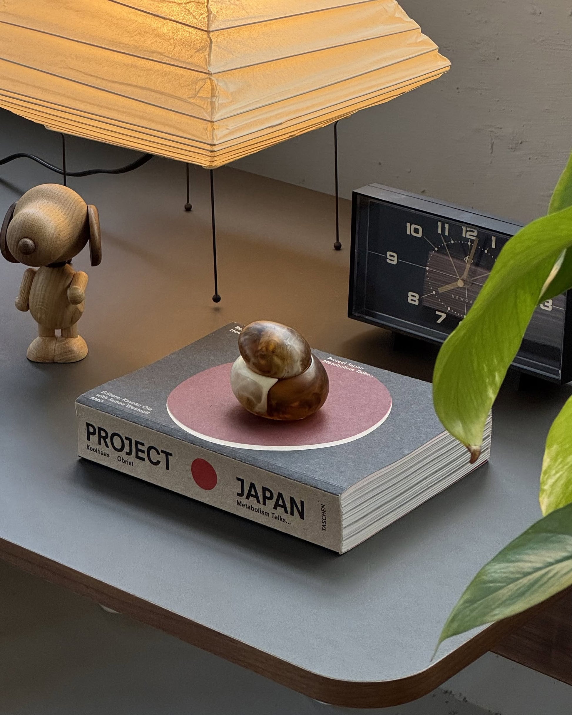 The Dinosaur Designs Small Pebble Jar, crafted from light brown wood with a sculptural design and matching lid beside it, is set against a plain, light beige background.