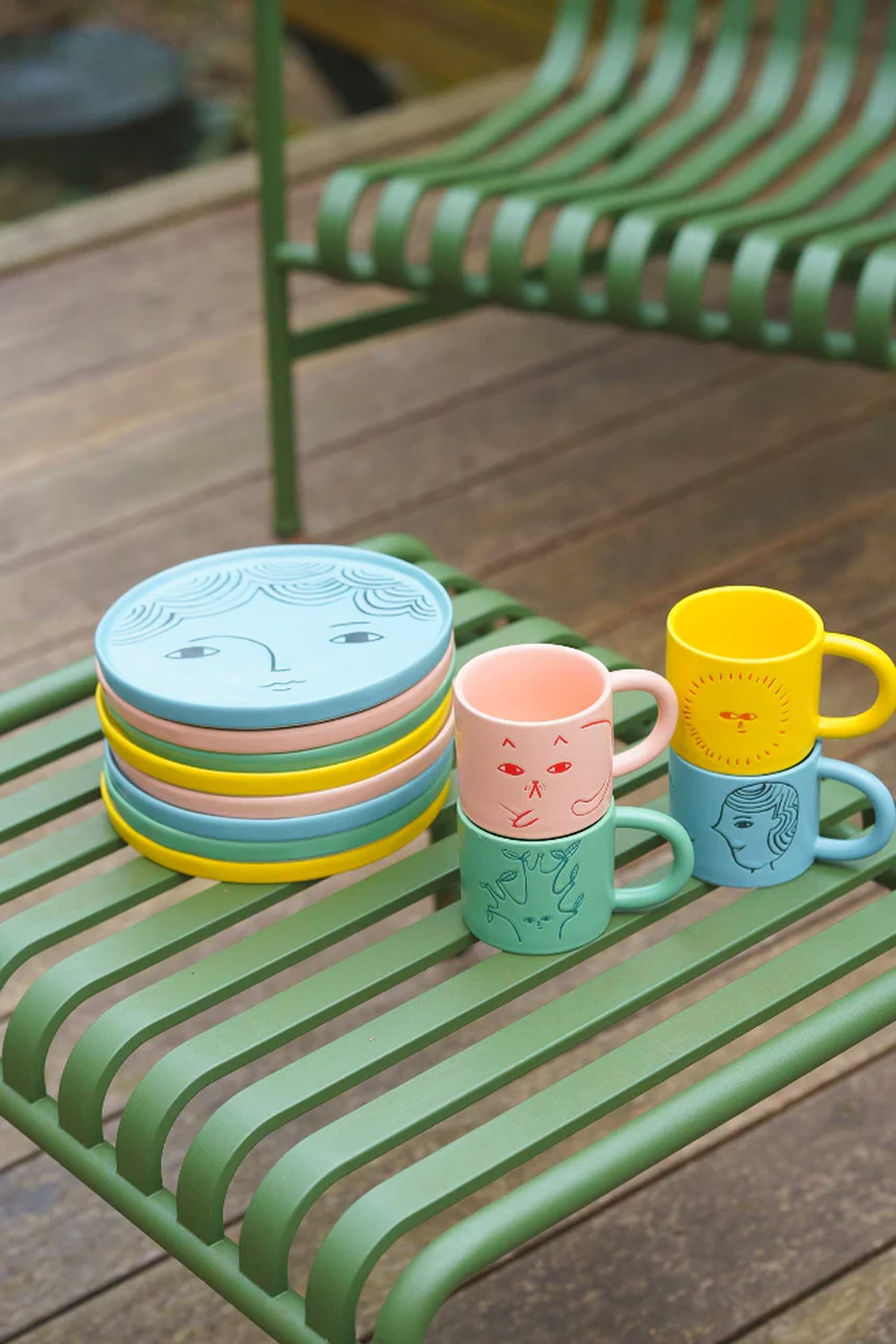 Four Donna Wilson Cat Side Plates and four matching mugs are stacked on a green metal chair with wooden decking in the background.