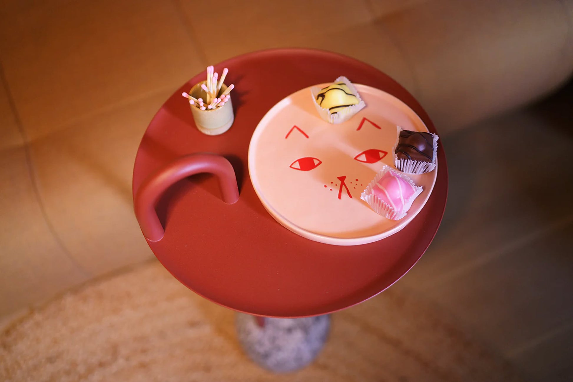 A small round red side table holds a Donna Wilson Cat Side Plate by Donna Wilson with three assorted pastries on top. Next to the plate is a cup of pink-tipped toothpicks. The table is placed beside a brown sofa.