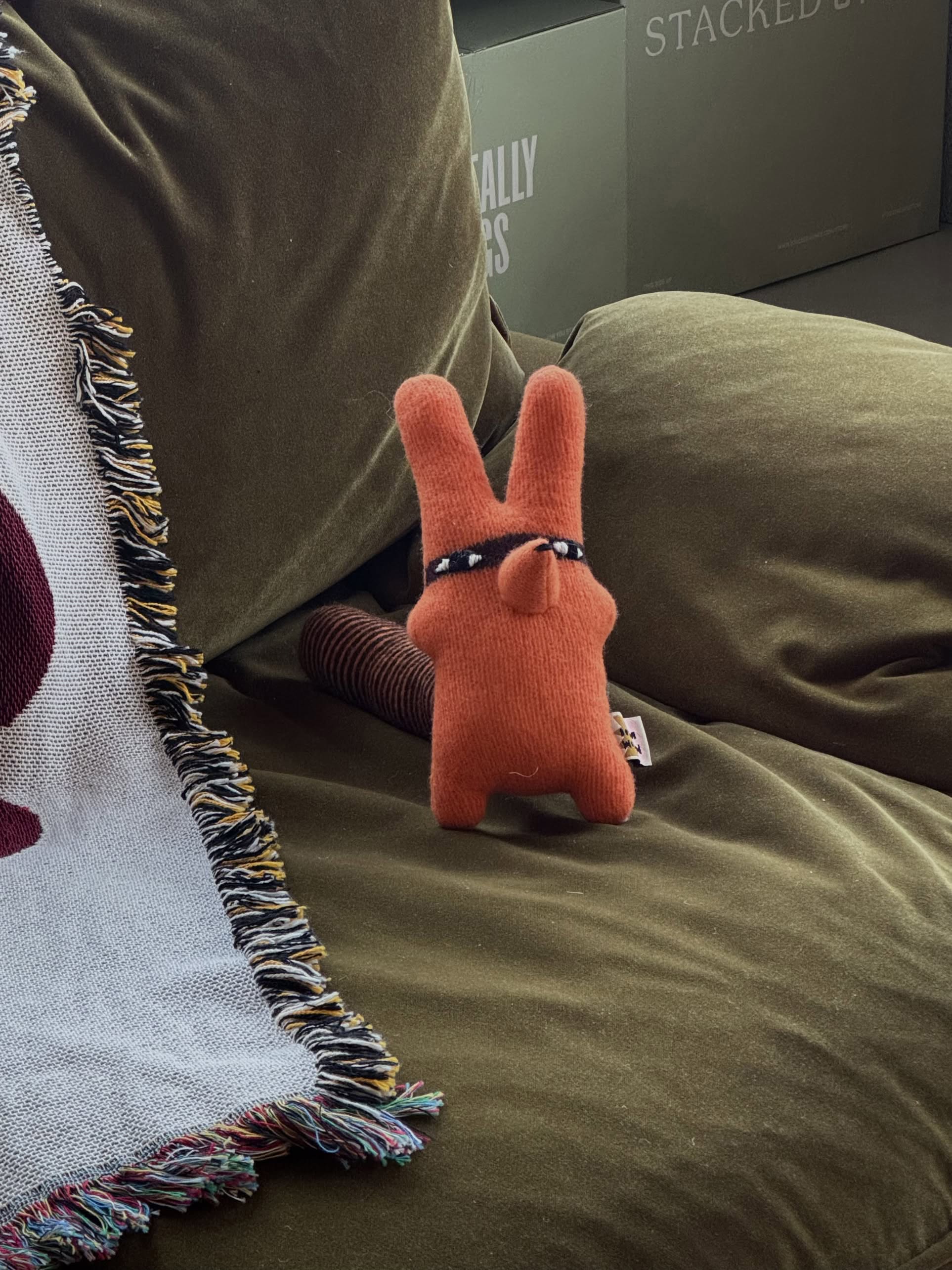 A Donna Wilson Creature - Rill, an orange lambswool plush toy with bunny ears, sits on an olive green couch beside a fringed blanket. Boxes with faint writing appear in the background.