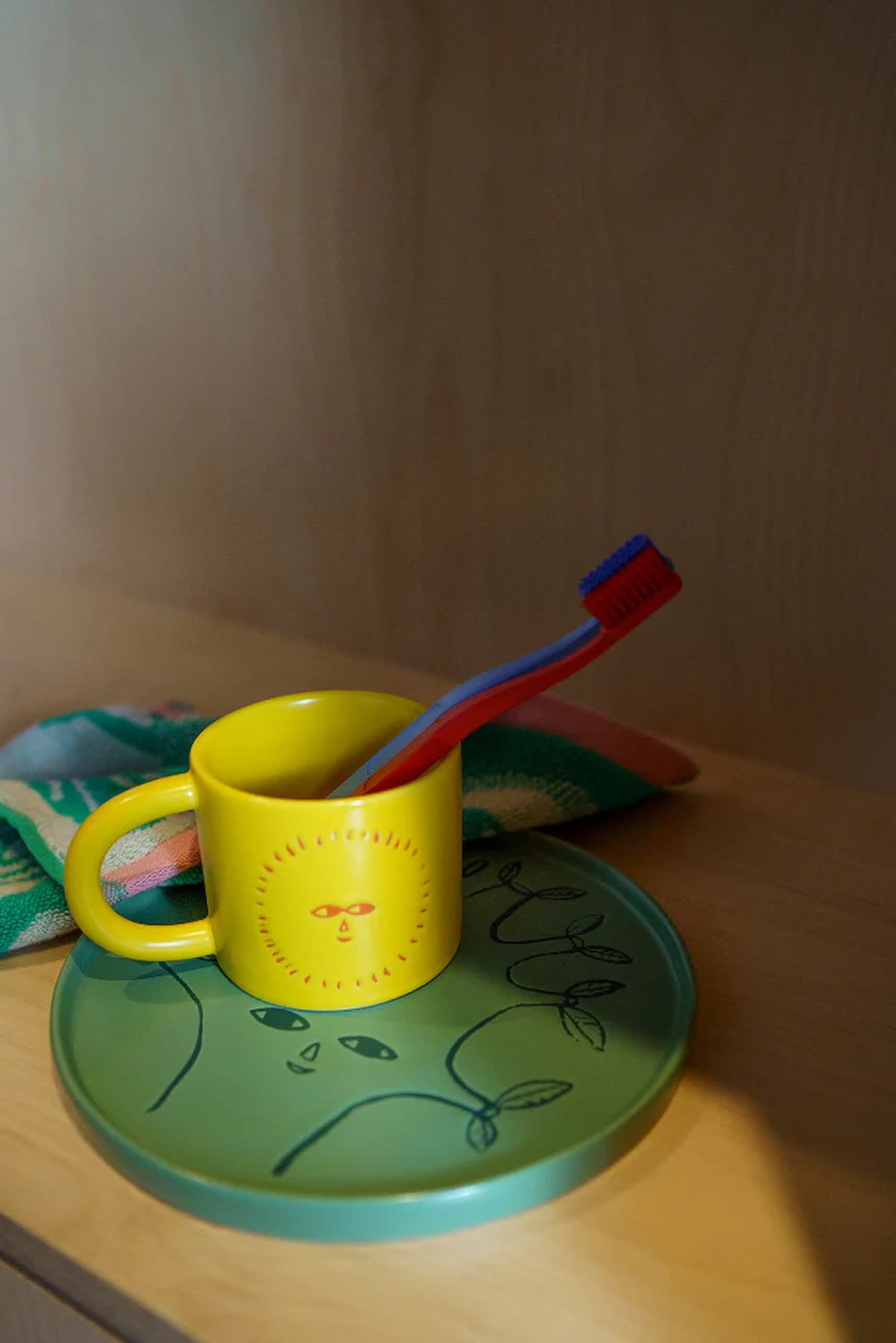 A yellow sun-face mug holds a red and blue toothbrush and sits on the Donna Wilson Tree Side Plate by Donna Wilson, alongside a folded green and pink towel—creating a unique display on a wooden surface.