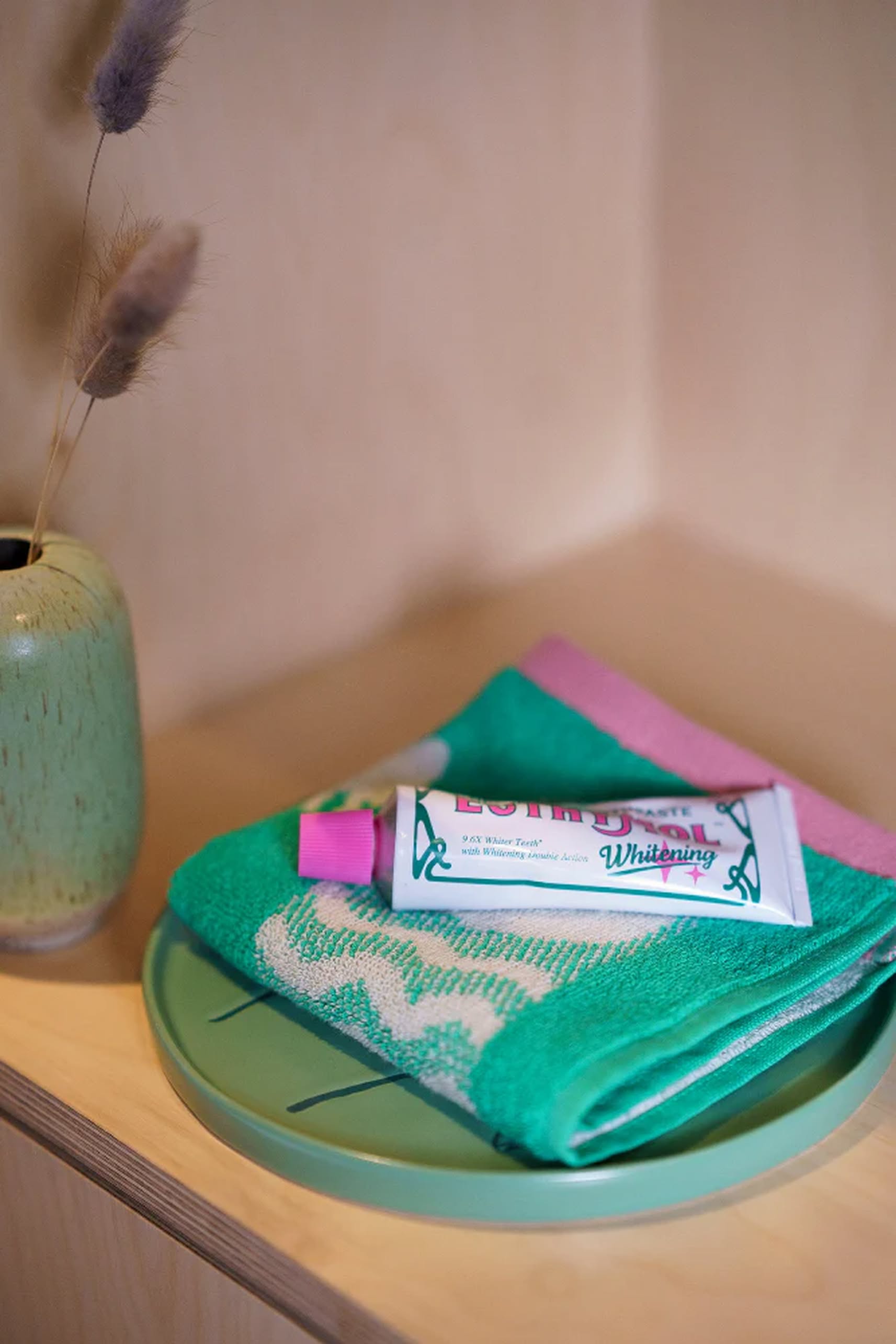 A green ceramic vase with dried grass sits beside a Donna Wilson Tree Side Plate by Donna Wilson, which holds a folded green towel and a tube of whitening toothpaste on a wooden surface.