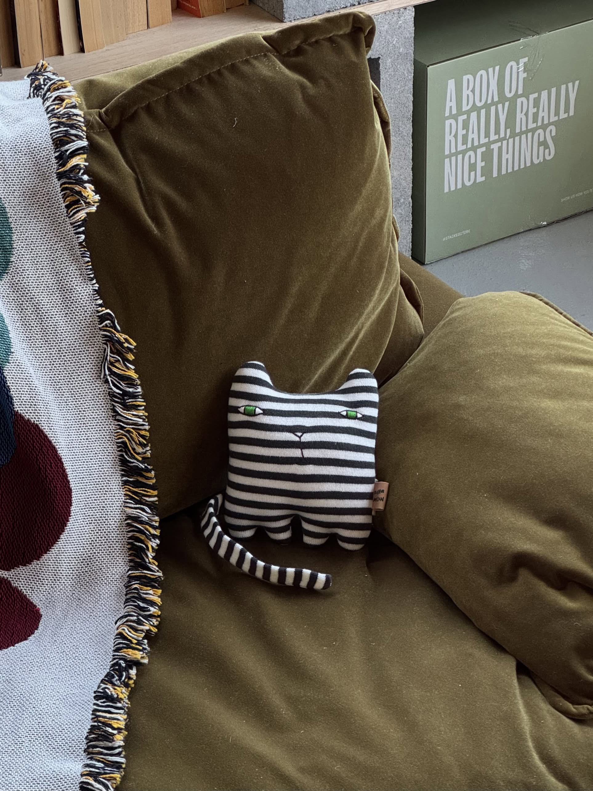 The Donna Wilson Wee One - Mono Kitten, a hand-embroidered plush cat with green eyes and organic cotton yarn by Donna Wilson, sits on an olive green couch beside a patterned blanket and a box reading A BOX OF REALLY, REALLY NICE THINGS.