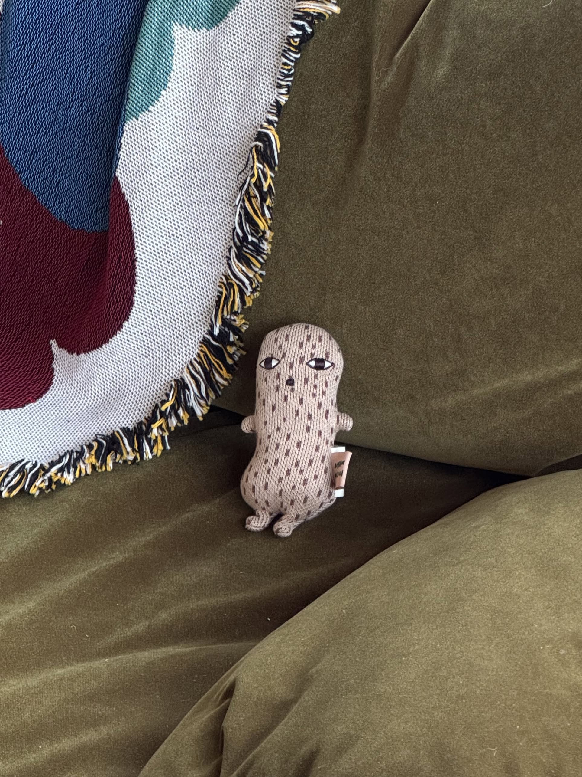 The Donna Wilson Wee One - Nutty, a small beige plush toy with a neutral expression and made from organic cotton, sits on an olive green couch beside a multicolored fringed woven blanket.