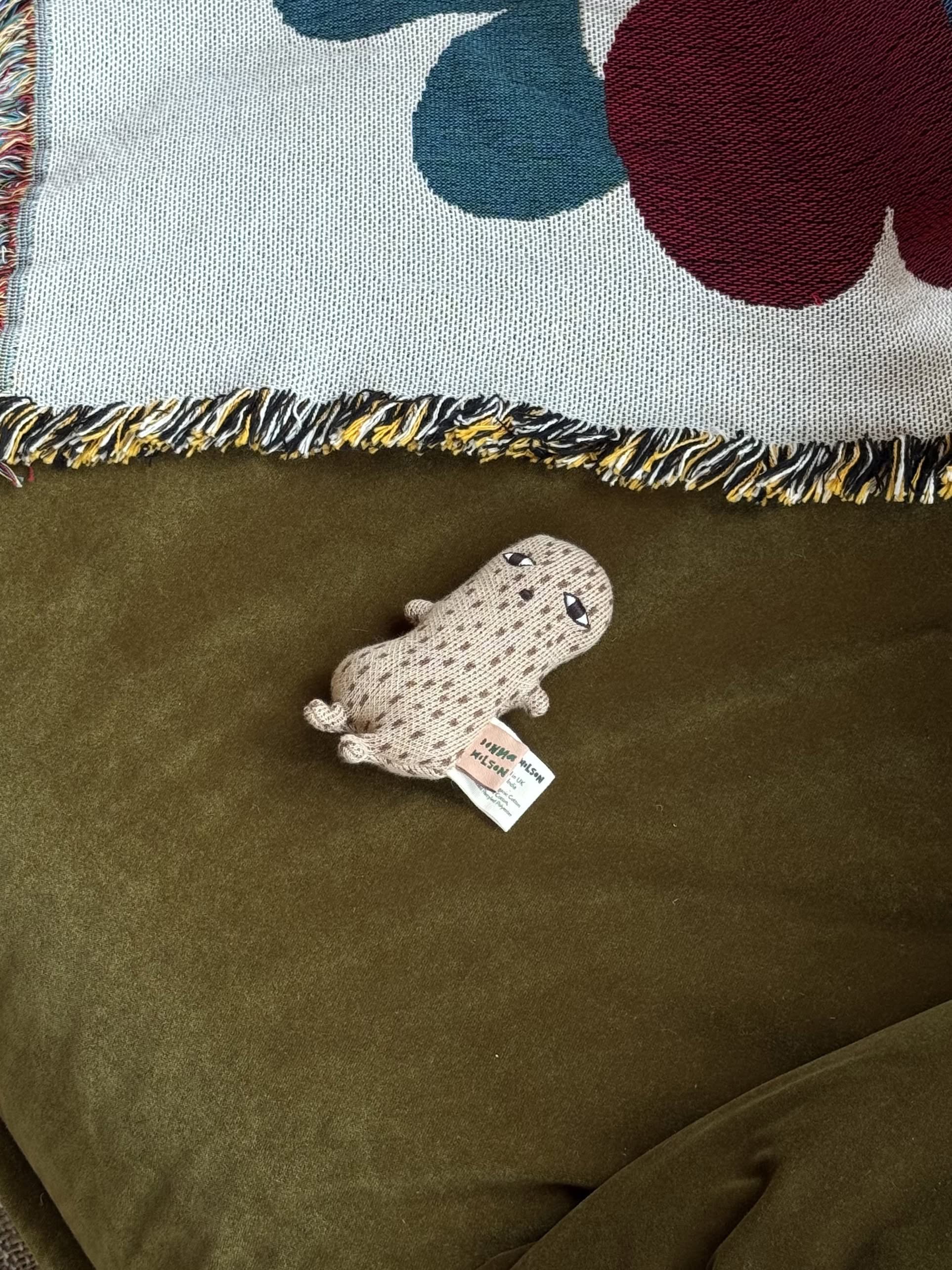 The Donna Wilson Wee One - Nutty, a small beige organic cotton plush toy with a drawn face, rests on an olive green velvet surface beside a fringed blanket with red and blue patterns.