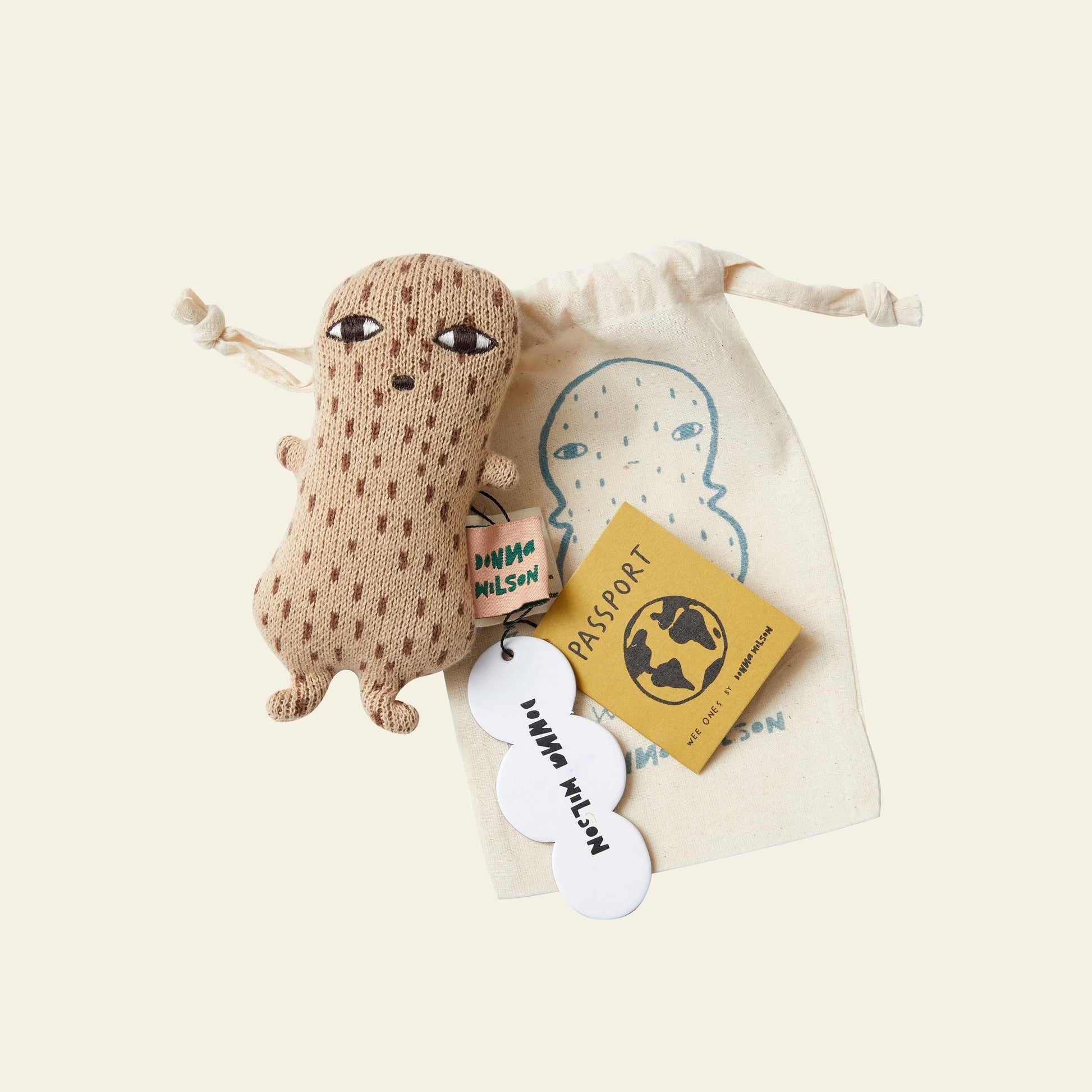 The Donna Wilson Wee One - Nutty, a hand-embroidered plush toy suitable from birth, comes with a mini yellow passport booklet and white tags, all placed on a beige drawstring bag featuring a blue outline of the toy.
