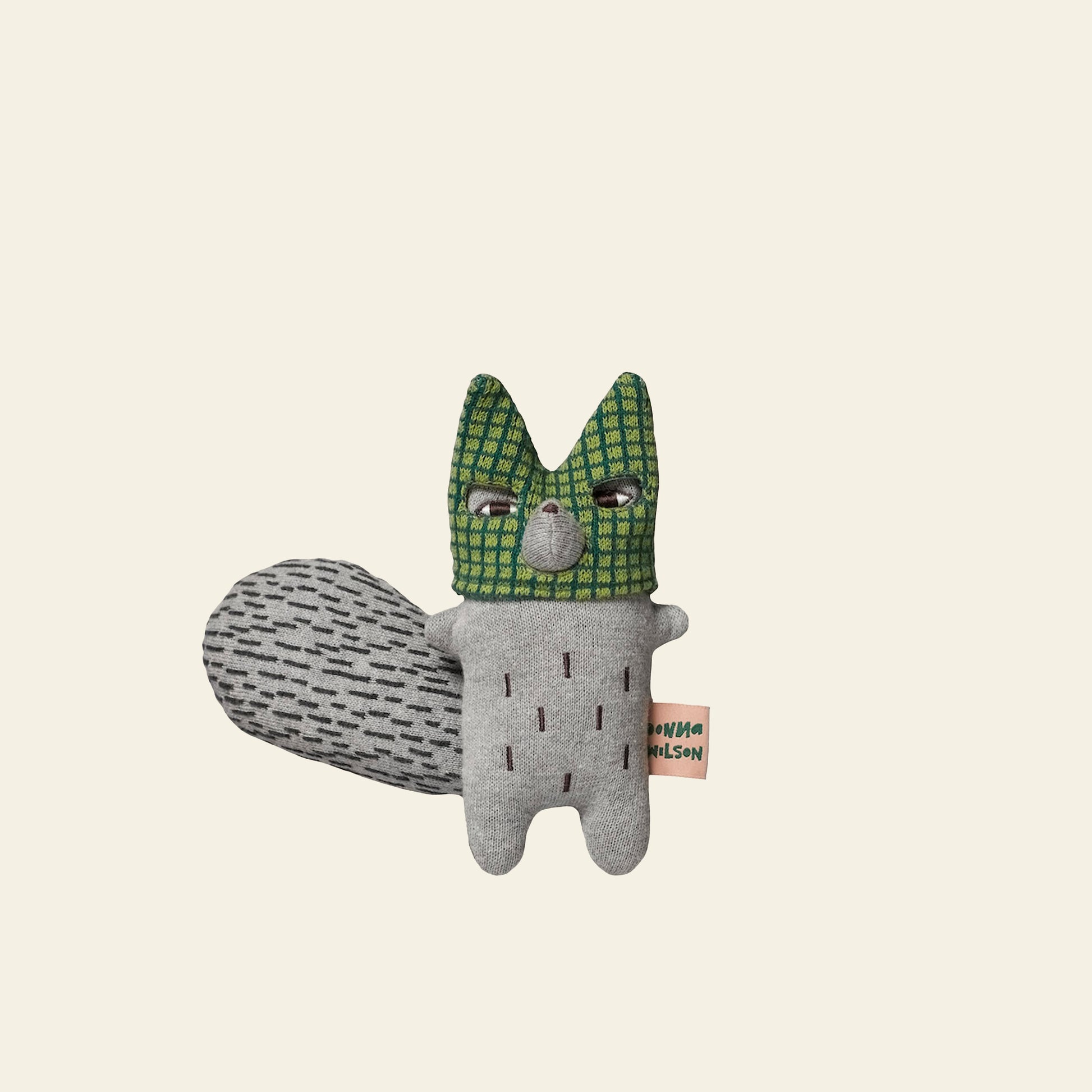 The Donna Wilson Wee One - Wilfred Wolf by Donna Wilson is a handmade plush toy crafted from organic cotton yarn, featuring a green checkered face, gray body, patterned tail, and hand-embroidered details. Shown on a beige background.