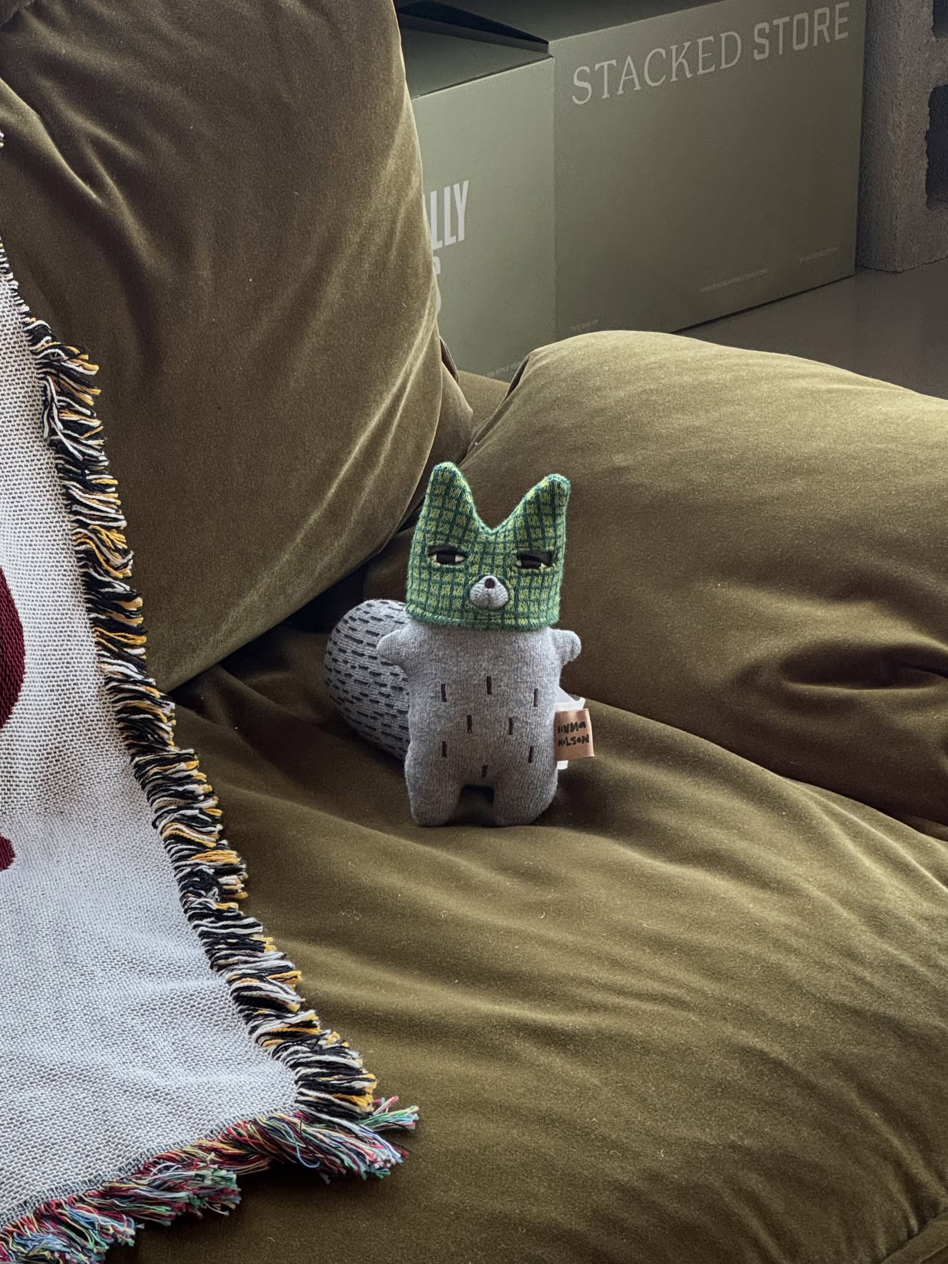 The Donna Wilson Wee One - Wilfred Wolf, a small plush toy by Donna Wilson, sits on an olive-green couch beside a colorful fringed blanket with cardboard boxes stacked in the background.