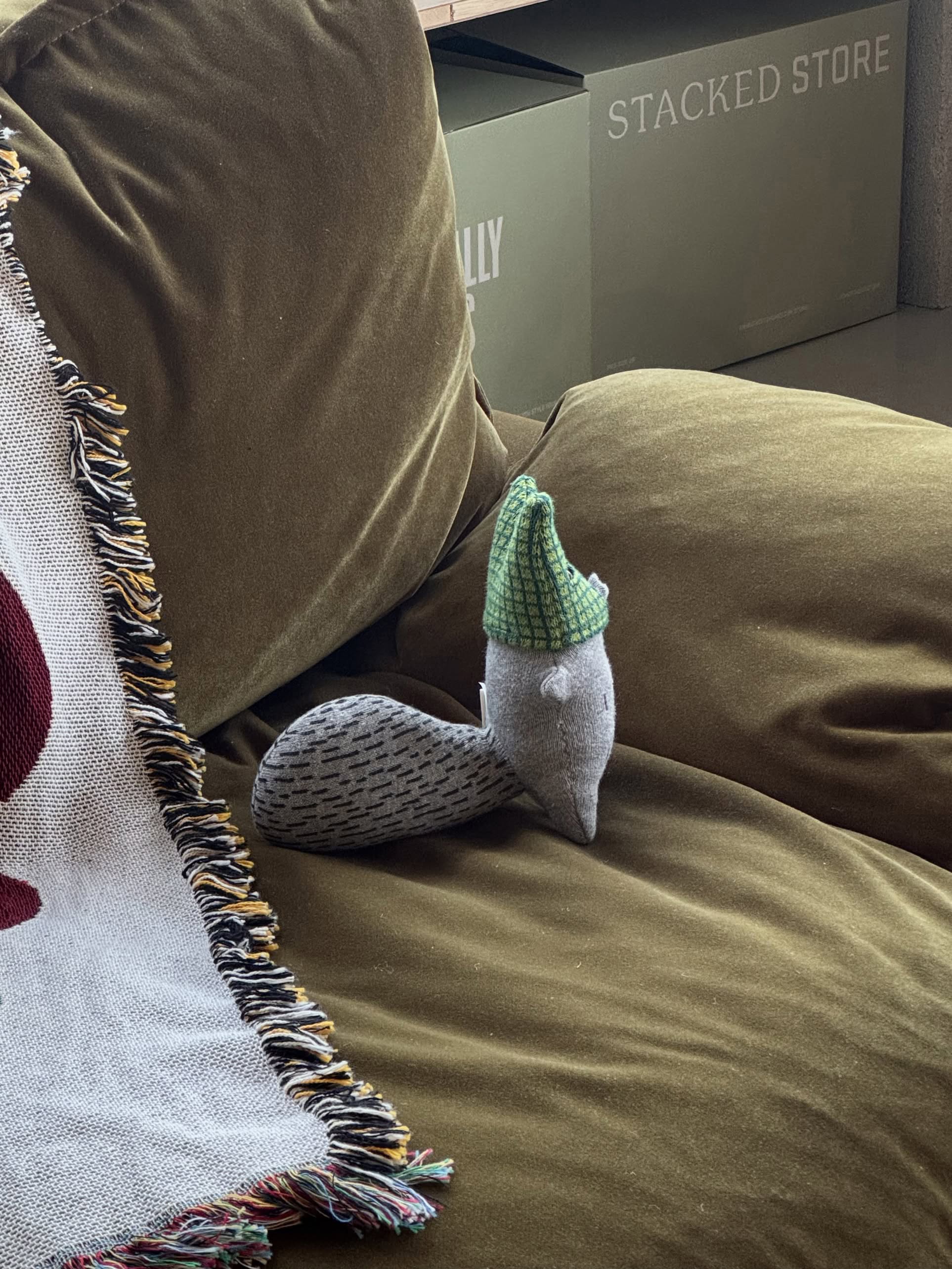 A Donna Wilson Wee One - Wilfred Wolf plush toy sits hand-embroidered on an olive-green couch beside a fringed blanket, while STACKED STORE green boxes are stacked against the wall in the background.