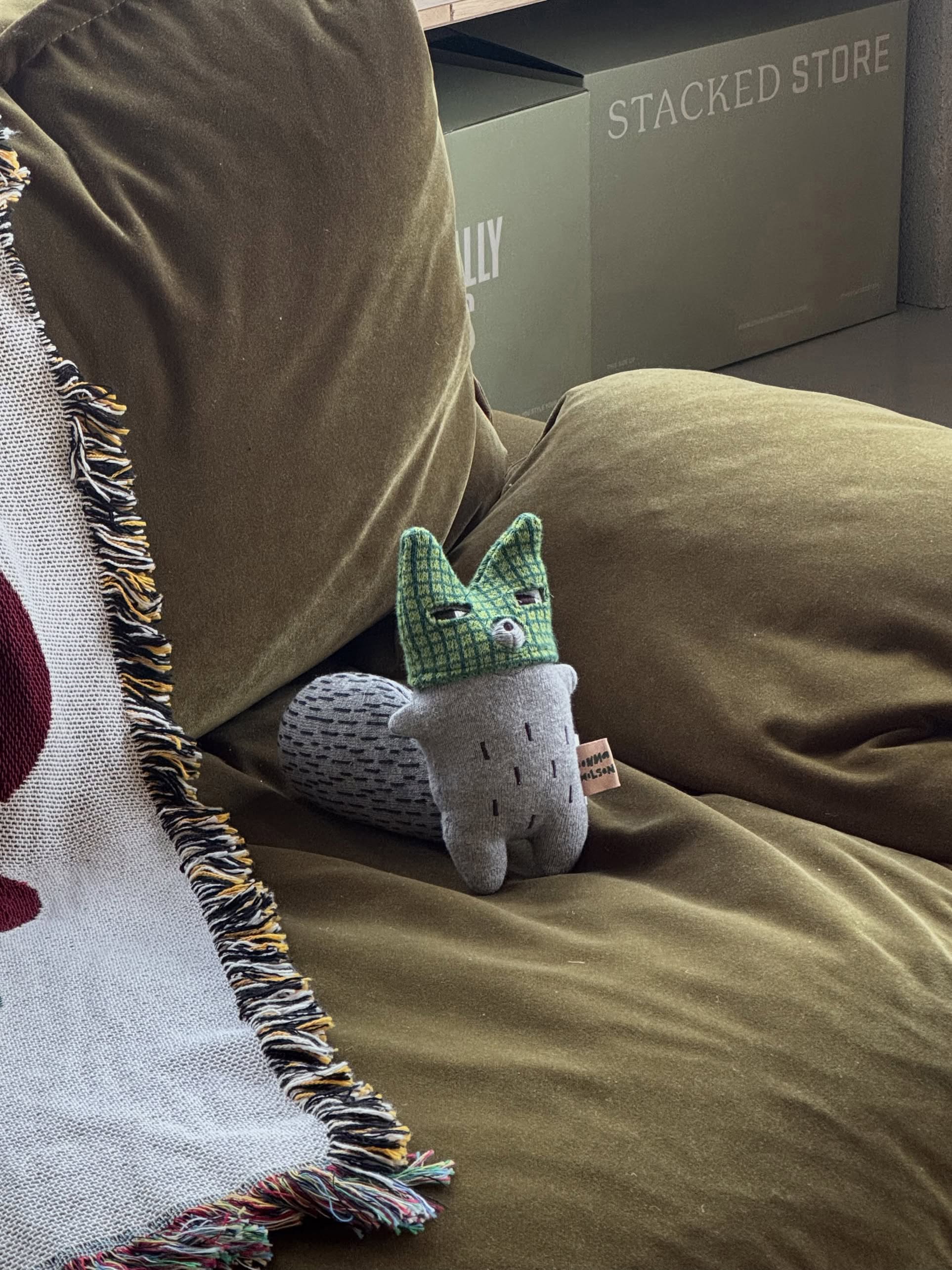 The Donna Wilson Wee One - Wilfred Wolf, a hand-embroidered organic cotton toy by Donna Wilson, sits on a green couch near a fringed blanket. Green STACKED STORE boxes are visible in the background.