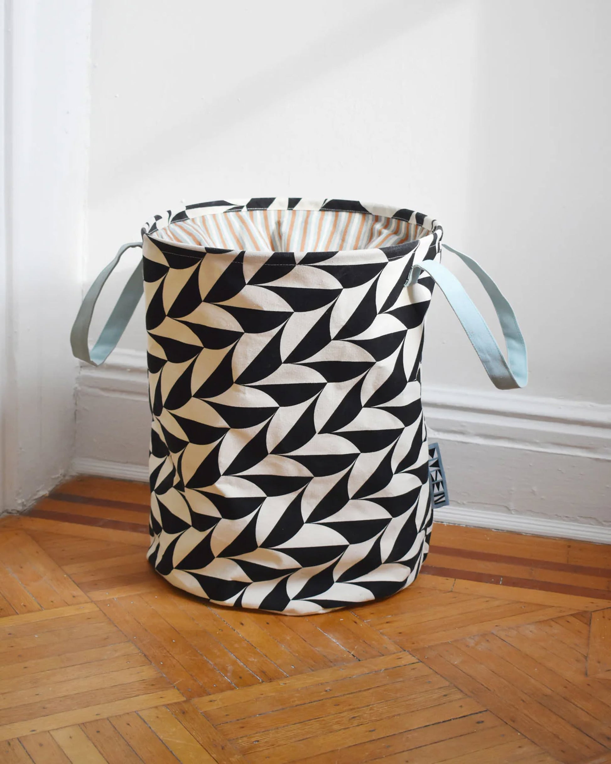 The Dusen Dusen Canvas Laundry Hamper by Dusen Dusen features a black and white geometric pattern, light blue handles, and an orange striped PE-coated interior. It sits on a wooden floor in the corner of a room.