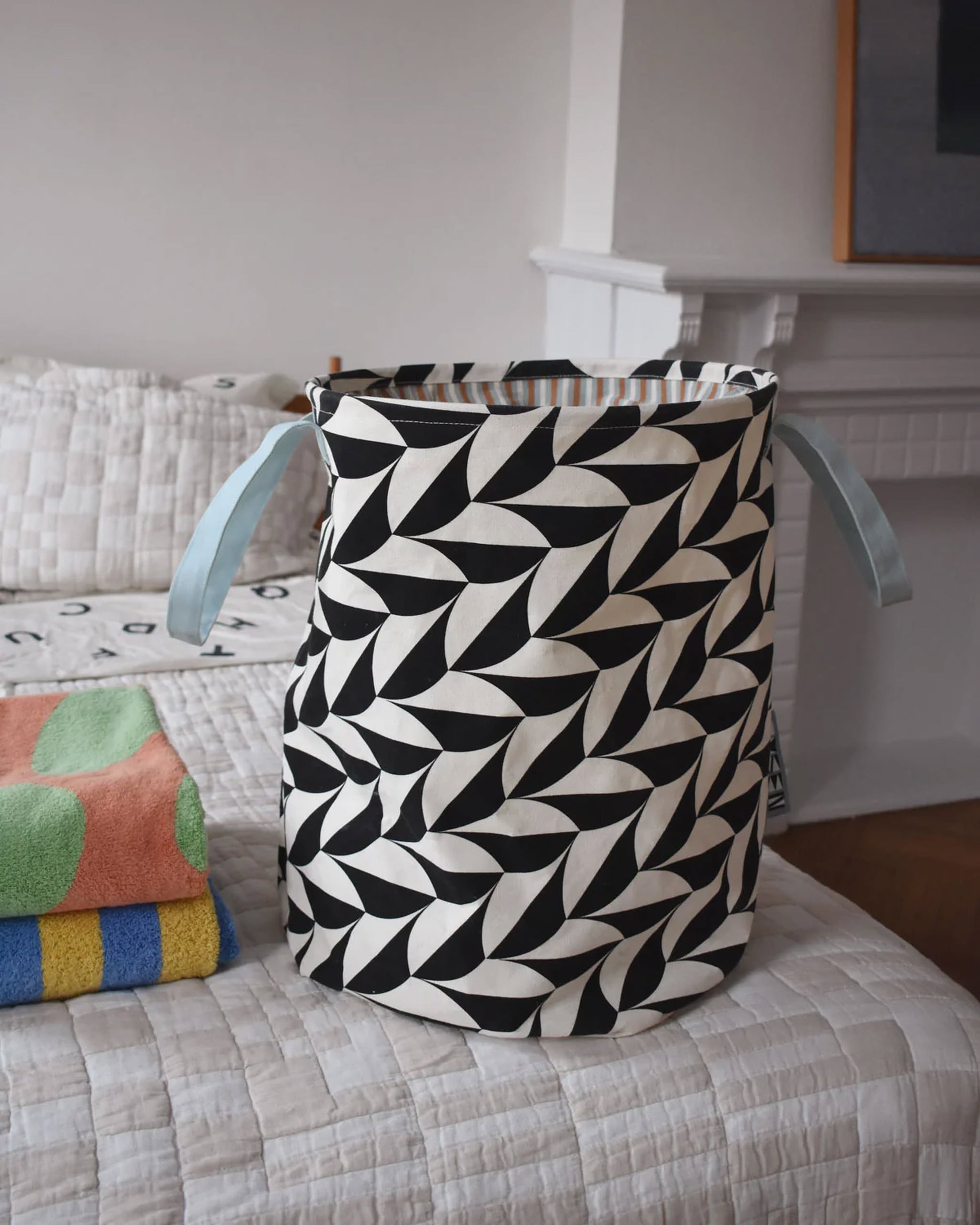 The Dusen Dusen Canvas Laundry Hamper by Dusen Dusen features a black and white scallop print, blue handles, and a durable PE-coated interior. It sits on a bed next to a folded towel, with a white fireplace and framed art in the background.