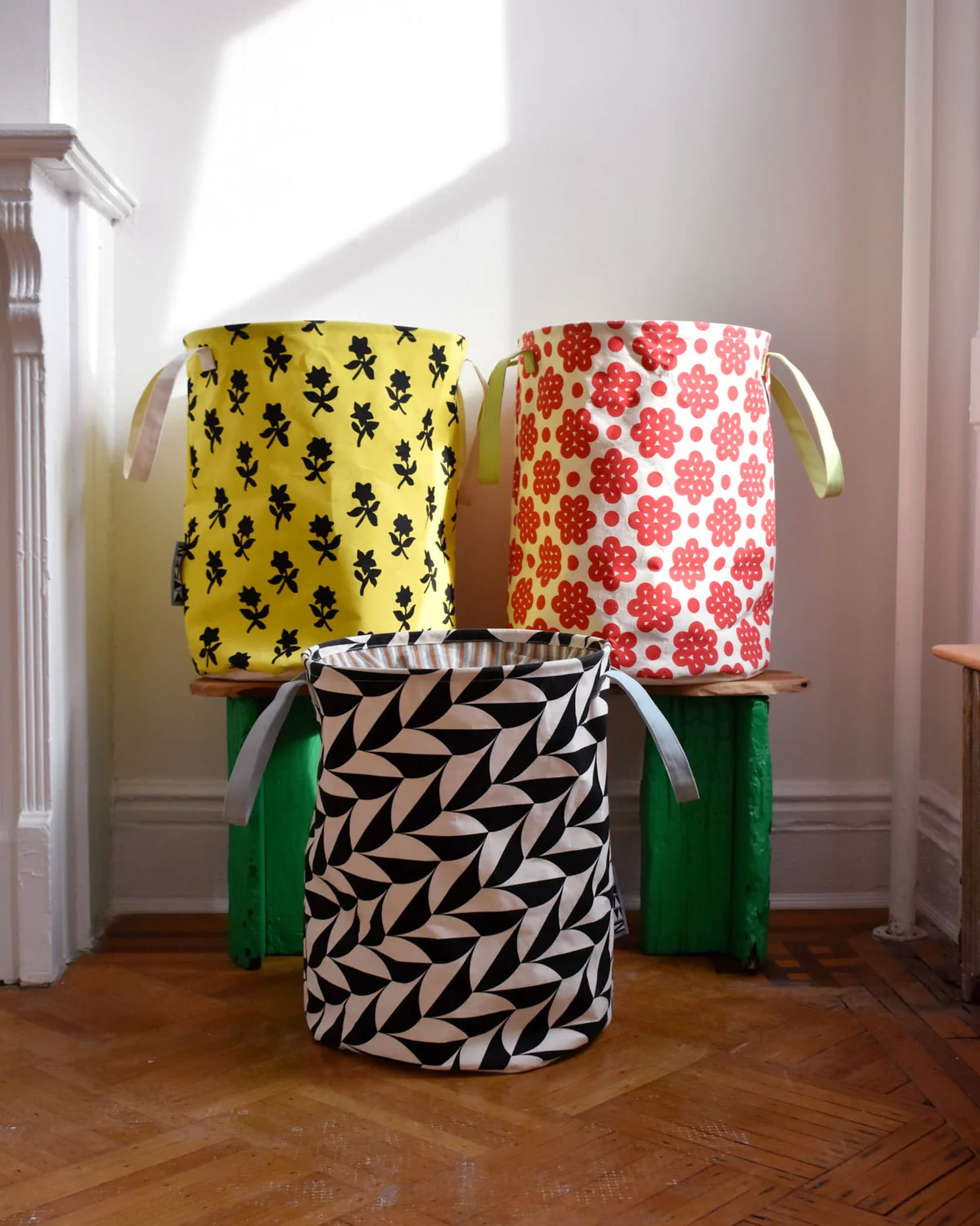 The Dusen Dusen Canvas Laundry Hamper set by Dusen Dusen features three handled bins—yellow with black flowers, red with white flowers, and black-and-white geometric designs—displayed on a wooden floor against a white wall.