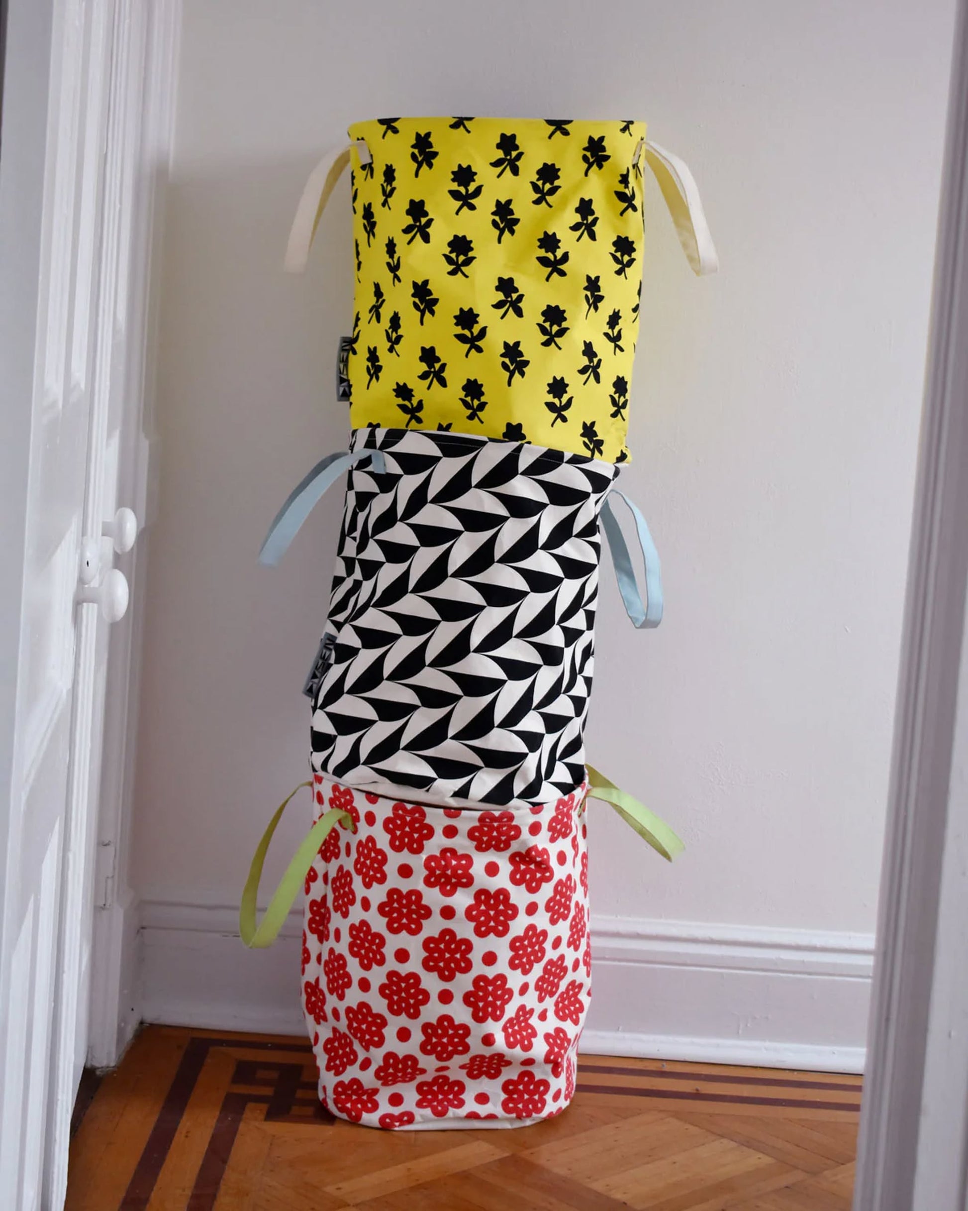 Three Dusen Dusen Canvas Laundry Hampers are stacked: the top has a yellow scallop print with black florals, the middle is black and white geometric, and the bottom is white with a red flower pattern.