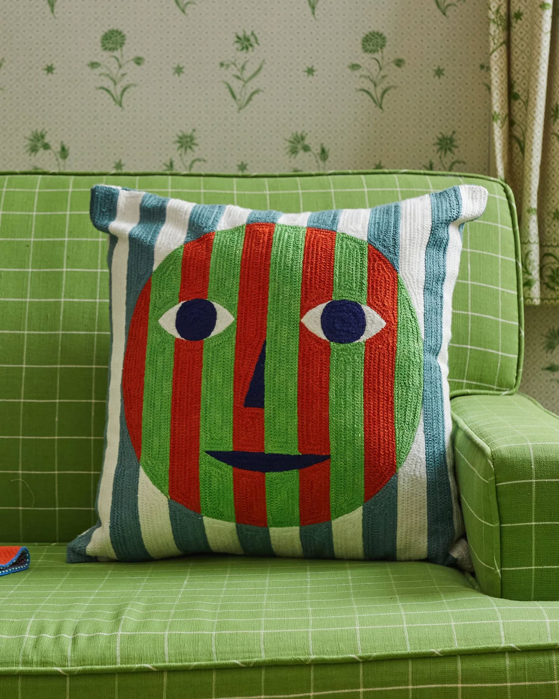 A green sofa with a white grid pattern showcases the Dusen Dusen Everybody Pillow Cover by Dusen Dusen, featuring a bold embroidered face with red and green stripes, blue and white eyes, and a simple smile on a striped background.