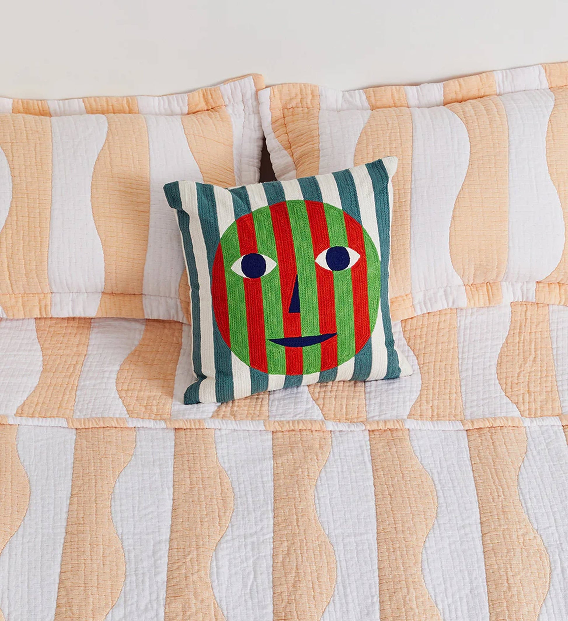 A bed with peach and white wavy bedding and a Dusen Dusen Everybody Pillow Cover—a striking accent by Dusen Dusen, showcasing a multi-coloured embroidered face with red and green stripes, blue-and-white eyes, and a smiling blue mouth.