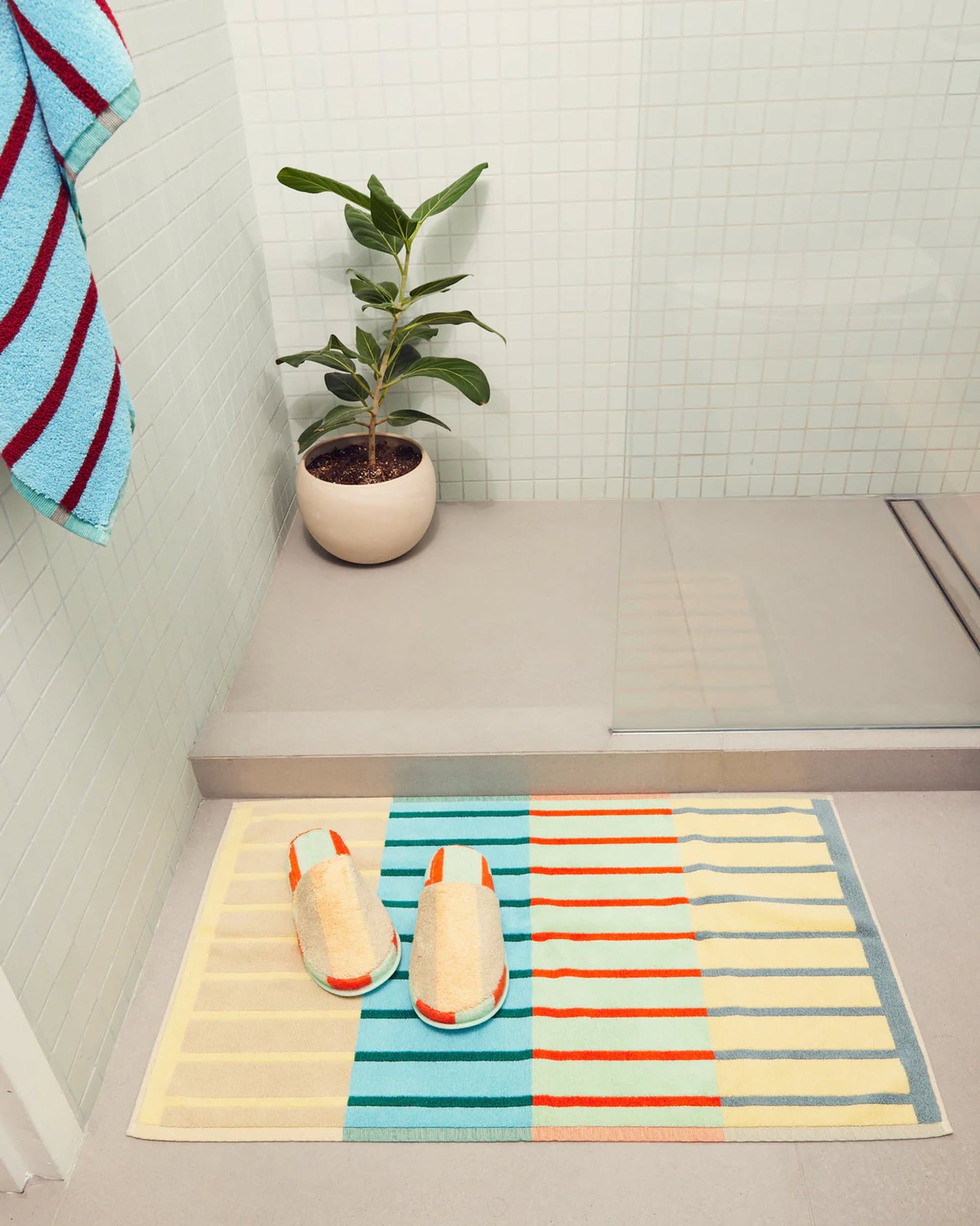 A bathroom scene with the Dusen Dusen House Stripe Concrete Bath Mat, matching striped slippers, a potted plant on the floor, a glass shower enclosure, and a striped towel. The palette is mostly pastel with pops of orange and blue.