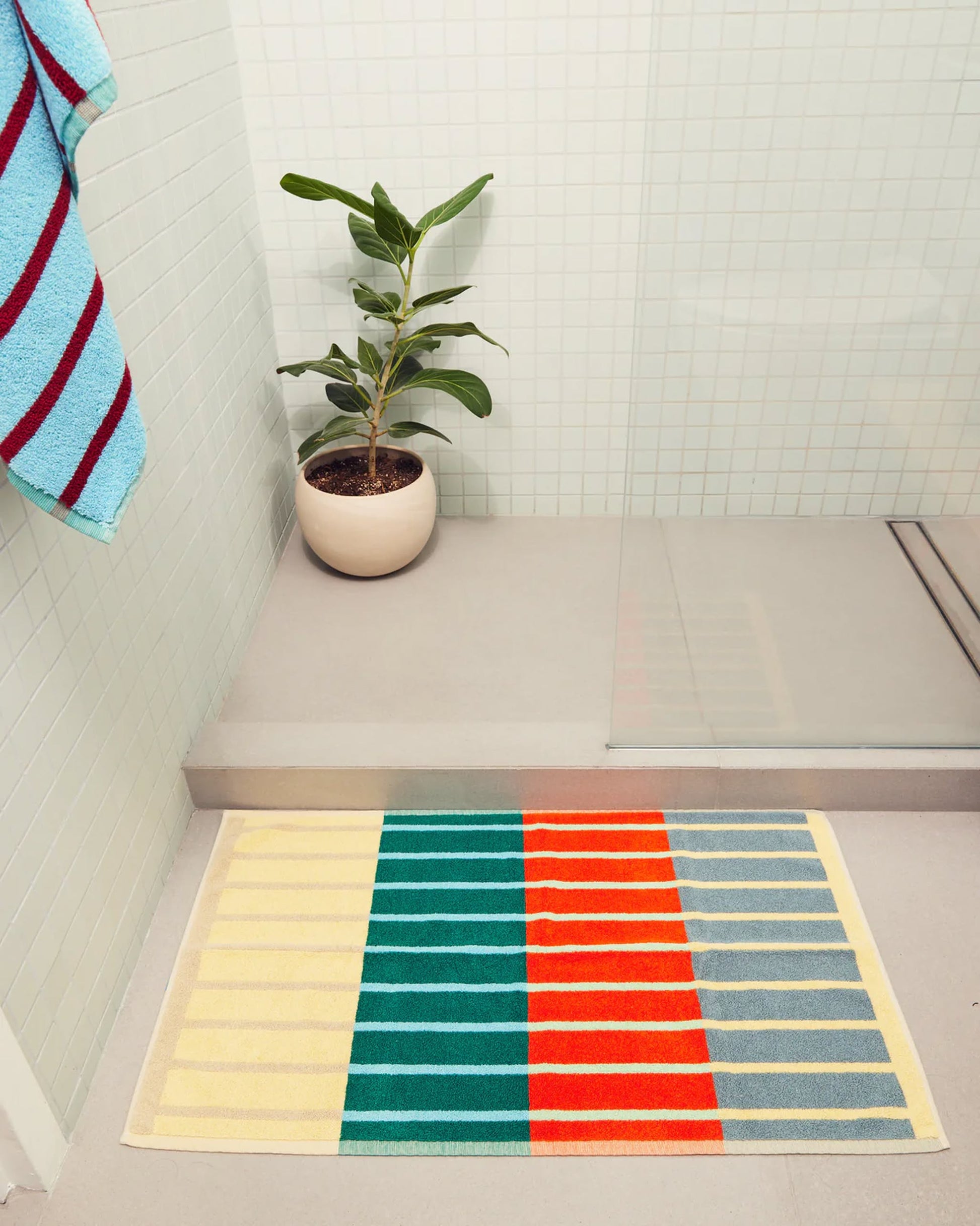 A bathroom with light tiles, a potted green plant, a Dusen Dusen House Stripe Concrete Bath Mat in yellow, green, orange, and gray stripes, and a multicolored towel hanging on the wall near a glass shower.