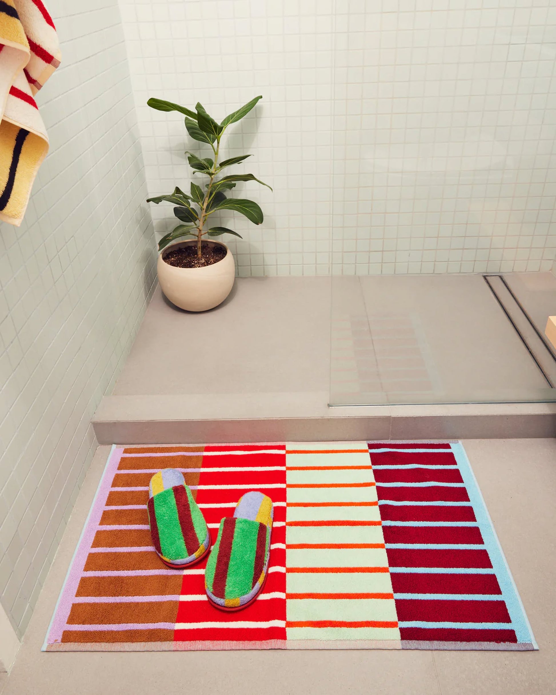 The Dusen Dusen House Stripe Copper Bath Mat, made from soft cotton terry, sits by a glass shower with matching slippers. A potted plant brightens the white tiled bathroom. Oeko-Tex Standard 100 certified for safe use.