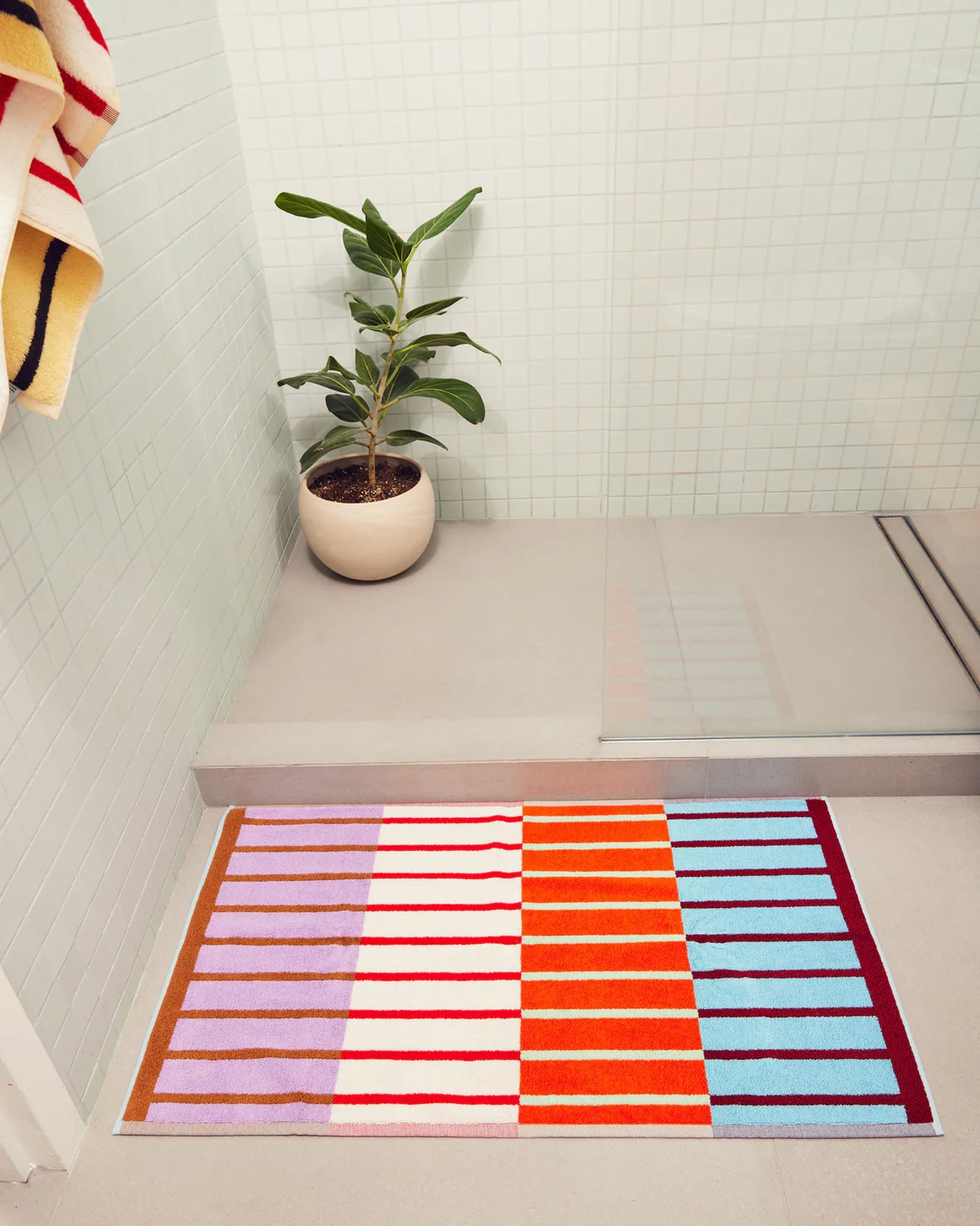 The Dusen Dusen House Stripe Copper Bath Mat by Dusen Dusen features colorful stripes in orange, blue, red, white, and lavender on soft Oeko-Tex Standard 100 certified cotton terry beside a potted plant and a glass-tiled shower.