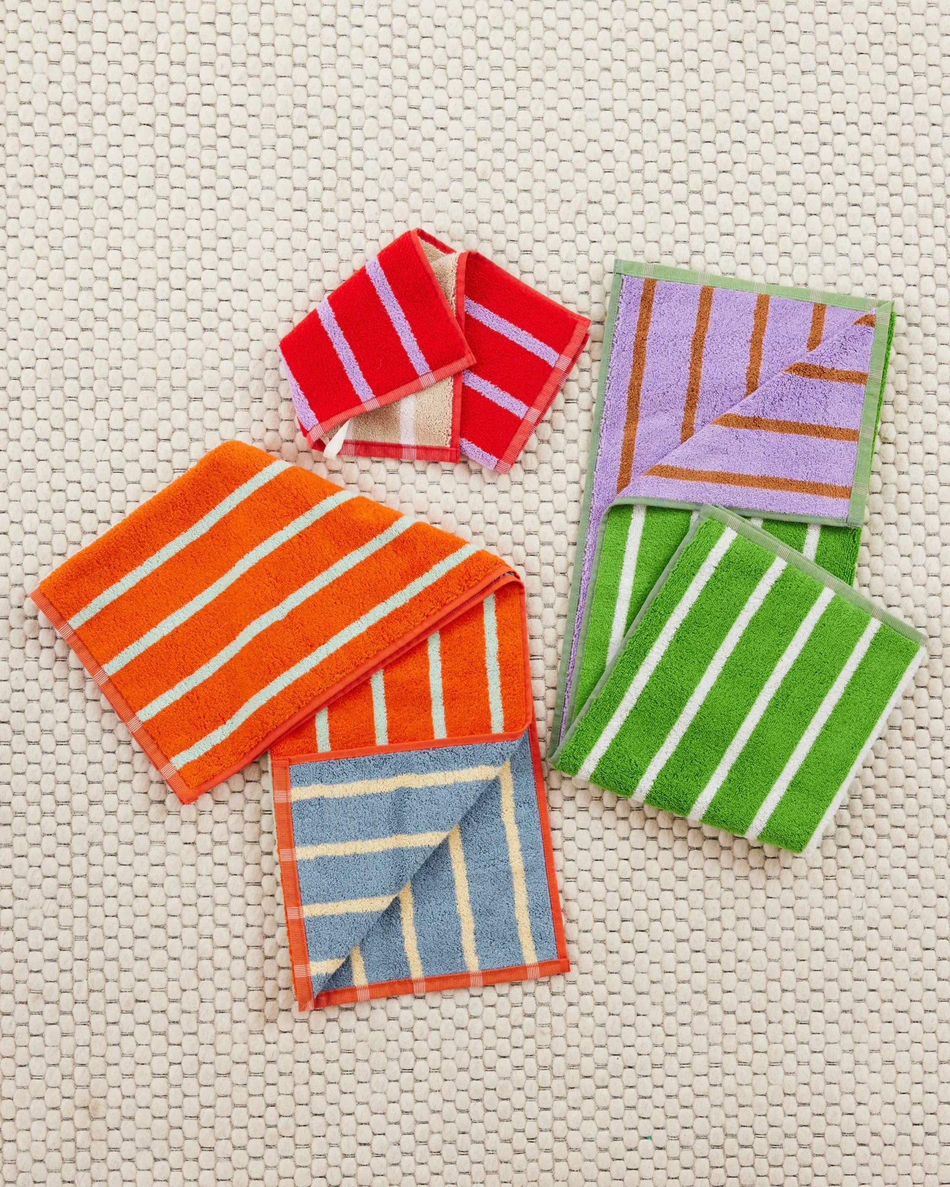 The Dusen Dusen House Stripe Porcelain Washcloth by Dusen Dusen, featuring vibrant red, orange, blue, green, and purple stripes in soft cotton terry, is displayed folded on a beige textured surface to show both sides.