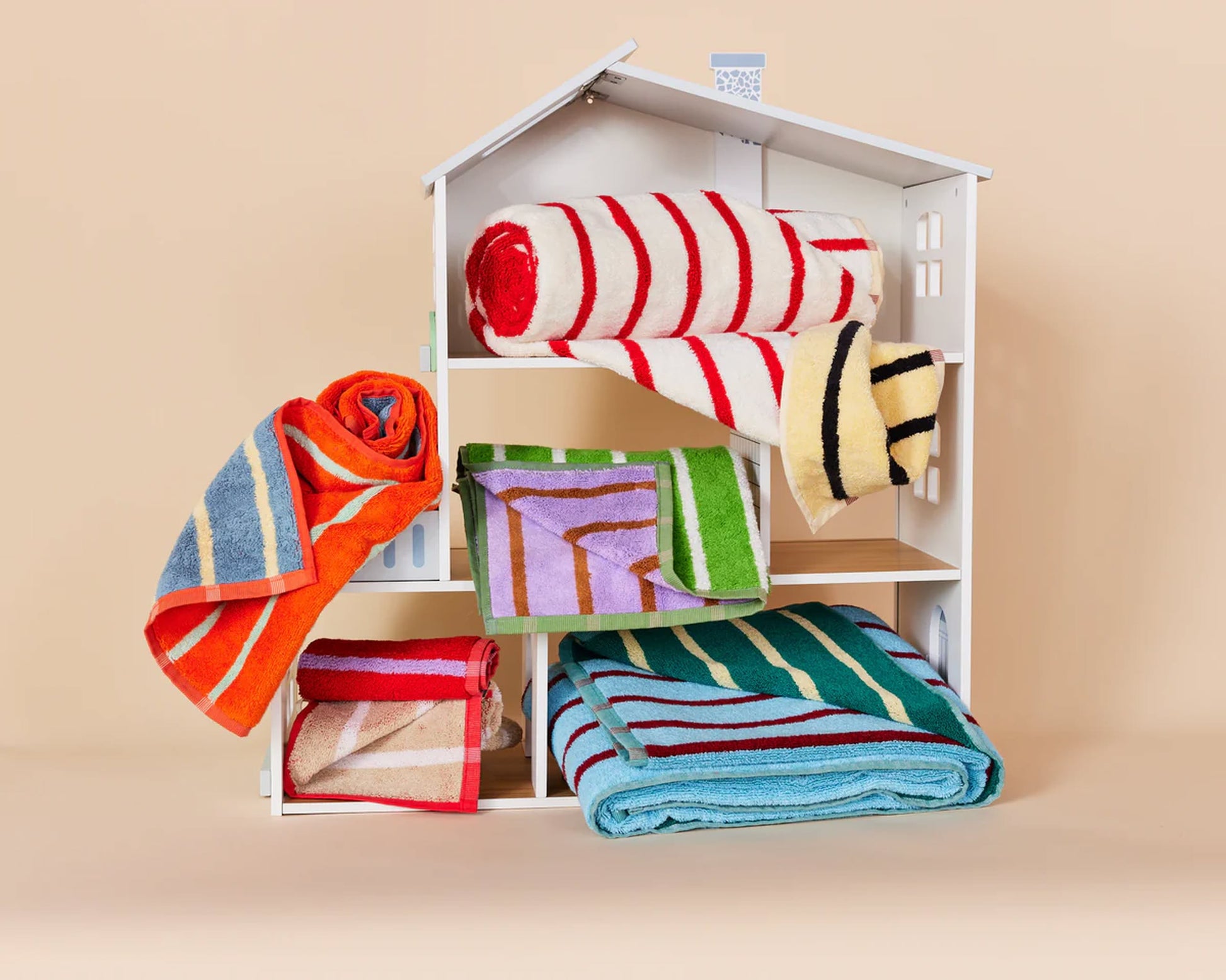 A small white dollhouse displays Dusen Dusen House Stripe Porcelain Washcloths with a vibrant striped pattern by Dusen Dusen, rolled and draped on its shelves against a light beige background.