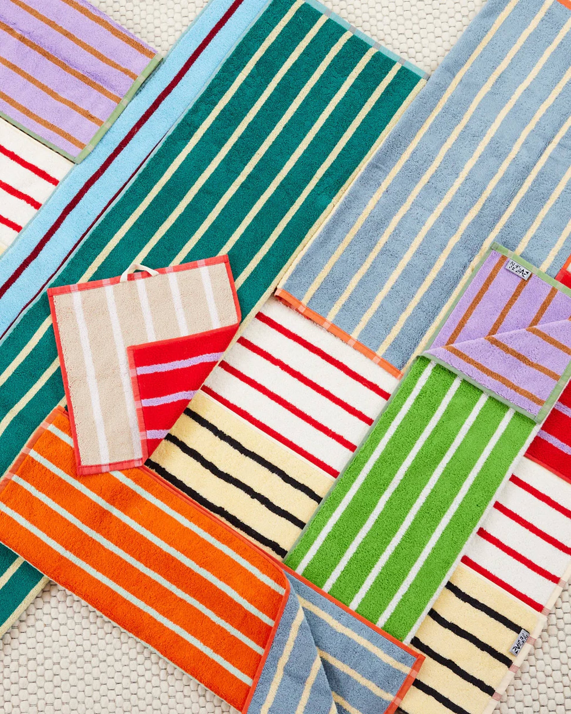 Dusen Dusen House Stripe Stone Hand Towels by Dusen Dusen feature vibrant green, blue, orange, red, purple, and beige stripes with geometric patterns and solid contrasting borders, displayed neatly on a woven off-white surface.