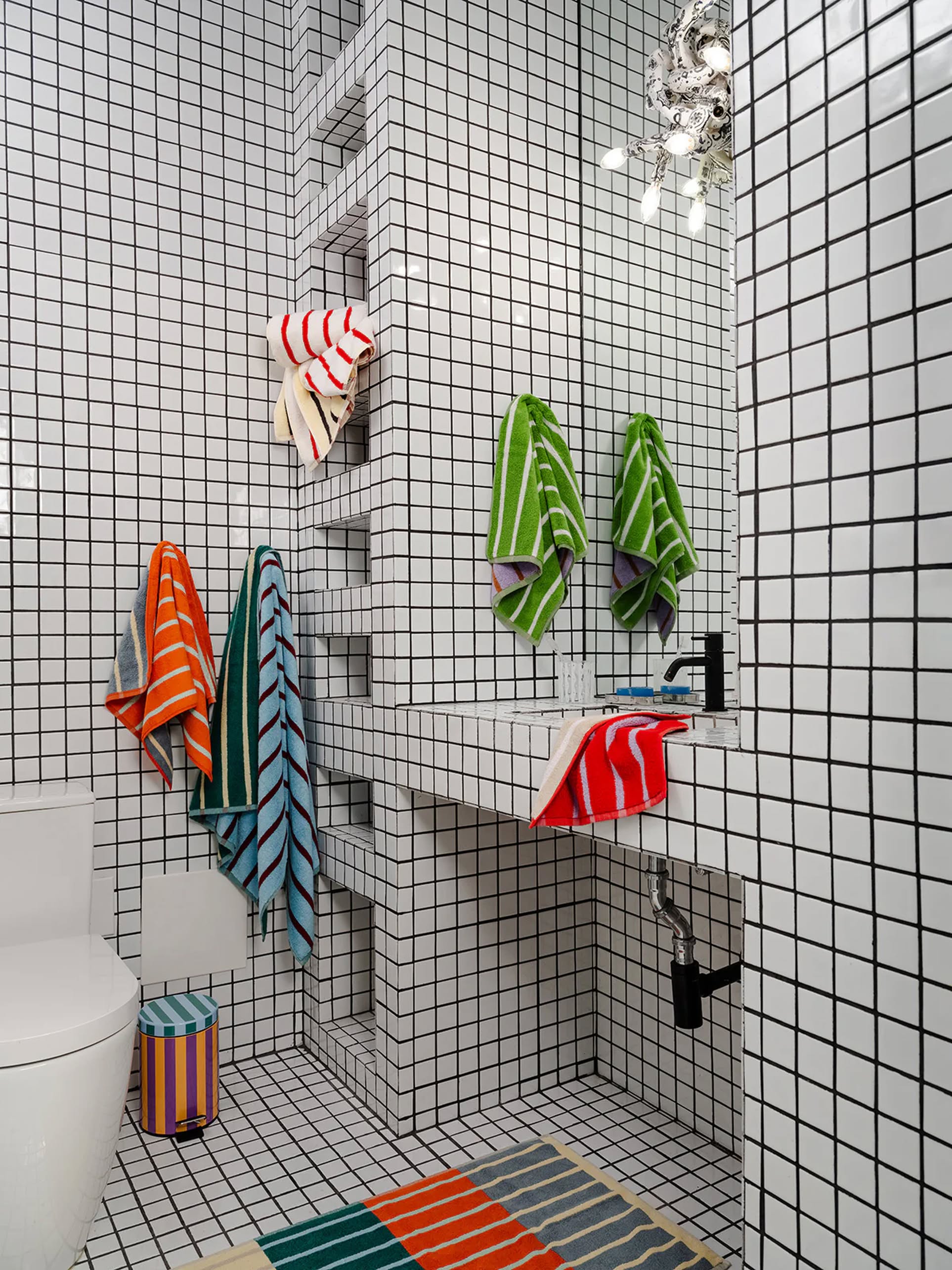 A bathroom with white square tiles and black grout features the Dusen Dusen House Stripe Stucco Bath Towel, showcasing bold stripes in vivid colors. A striped rug, multicolored bin, and modern white fixtures complete the lively look.