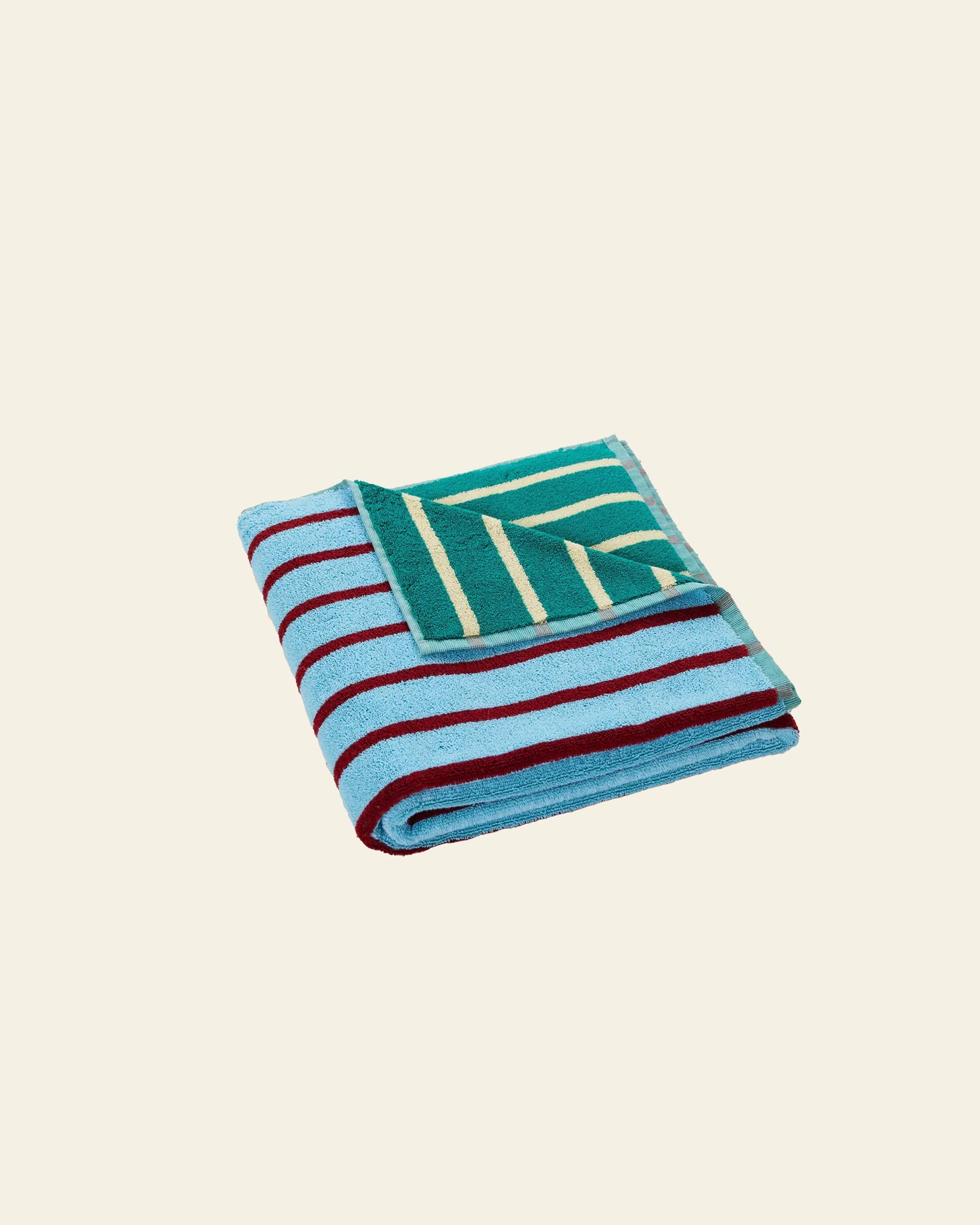 The Dusen Dusen House Stripe Tile Bath Towel by Dusen Dusen features bold blue and burgundy stripes on cotton terry, accented by a vibrant green corner with yellow diagonal stripes, set against a plain cream background.