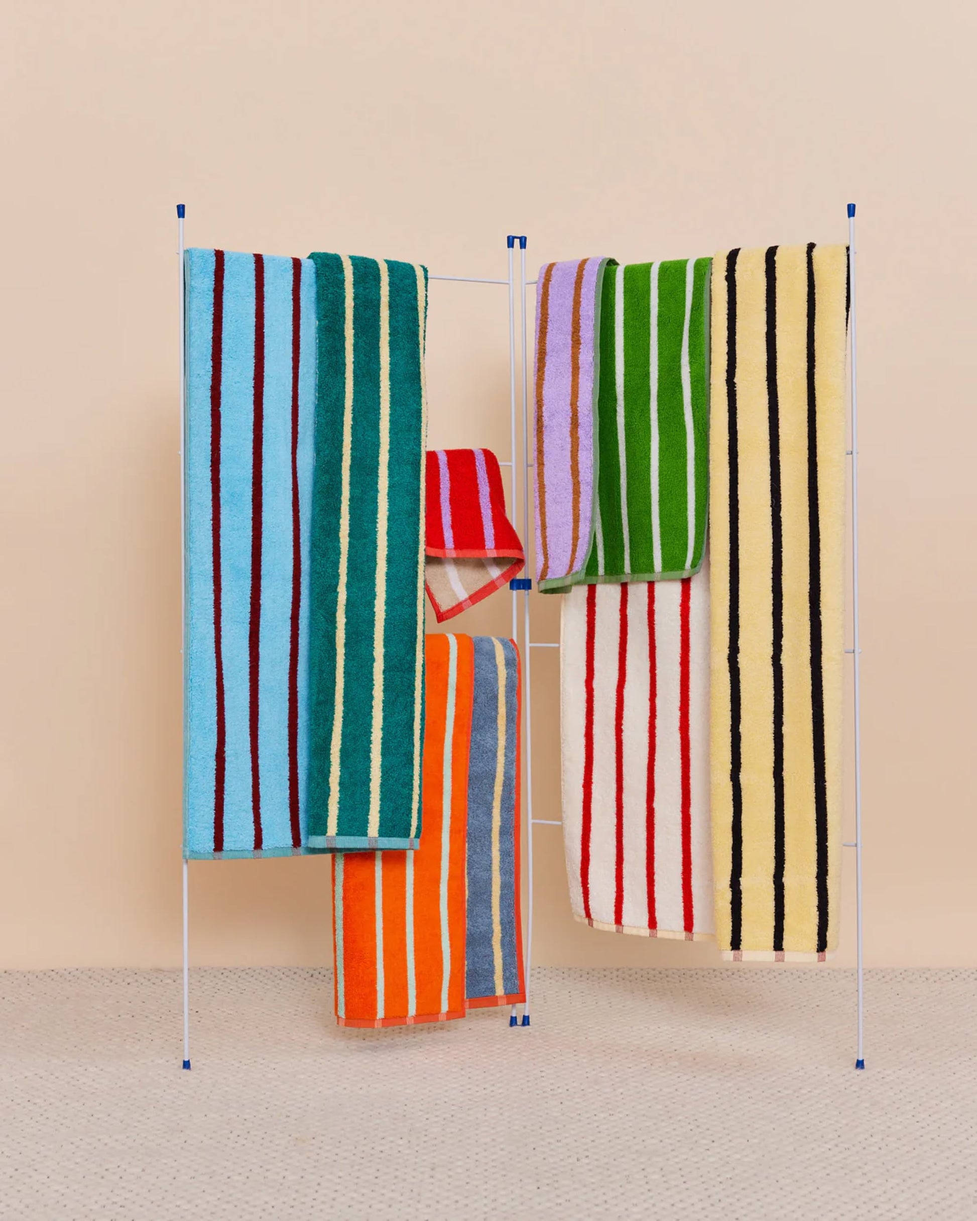 A standing drying rack holds five Dusen Dusen House Stripe Tile Bath Towels, featuring bold cotton terry stripes in green, blue, orange, yellow, red, and purple by Dusen Dusen on a beige background and light gray patterned floor.