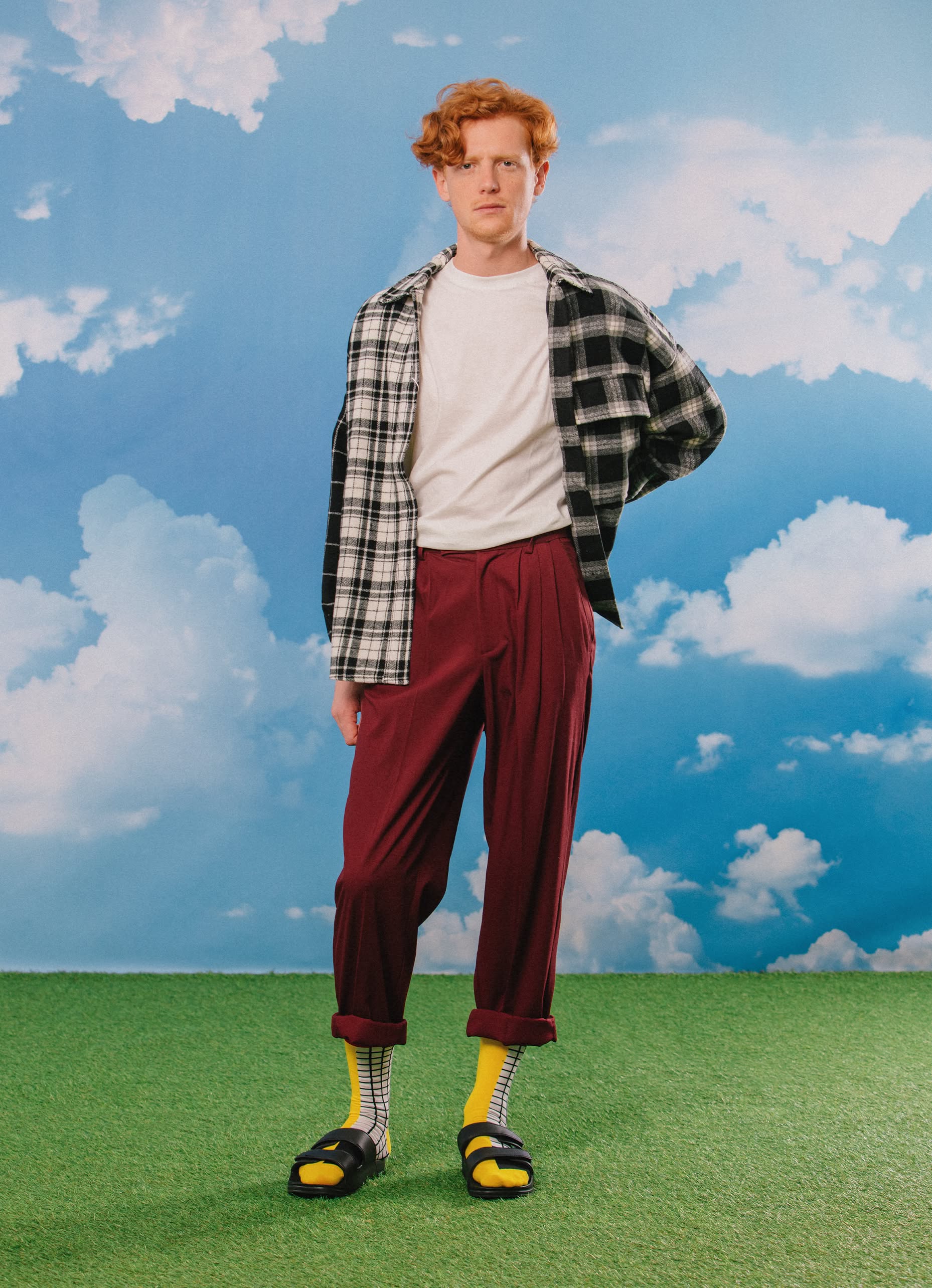 A person with short red hair stands on green grass under a blue sky, wearing a white T-shirt, black-and-white plaid overshirt, maroon pants, black sandals, and Eat My Socks French Fries Socks by Eat My Socks for a quirky accessories touch.