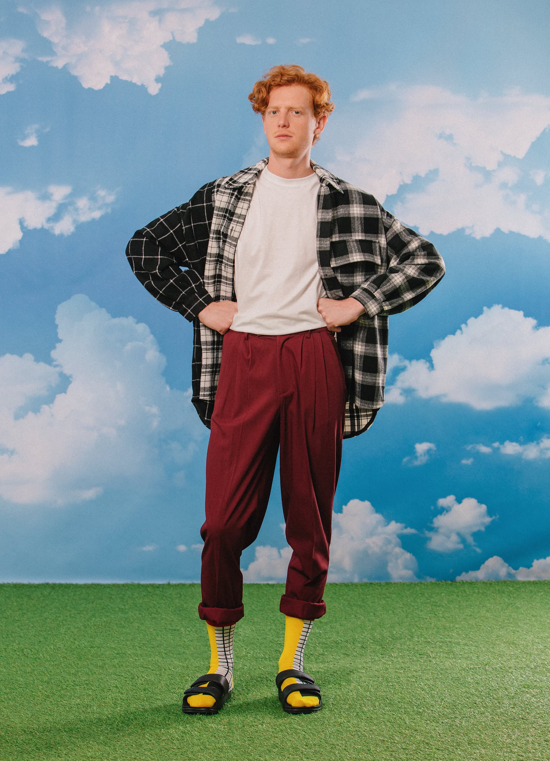 A red-haired person stands on green grass under a blue sky, wearing a white tee, plaid overshirt, maroon pants, black sandals, and Eat My Socks French Fries Socks by Eat My Socks—showing off quirky style with unique accessories.