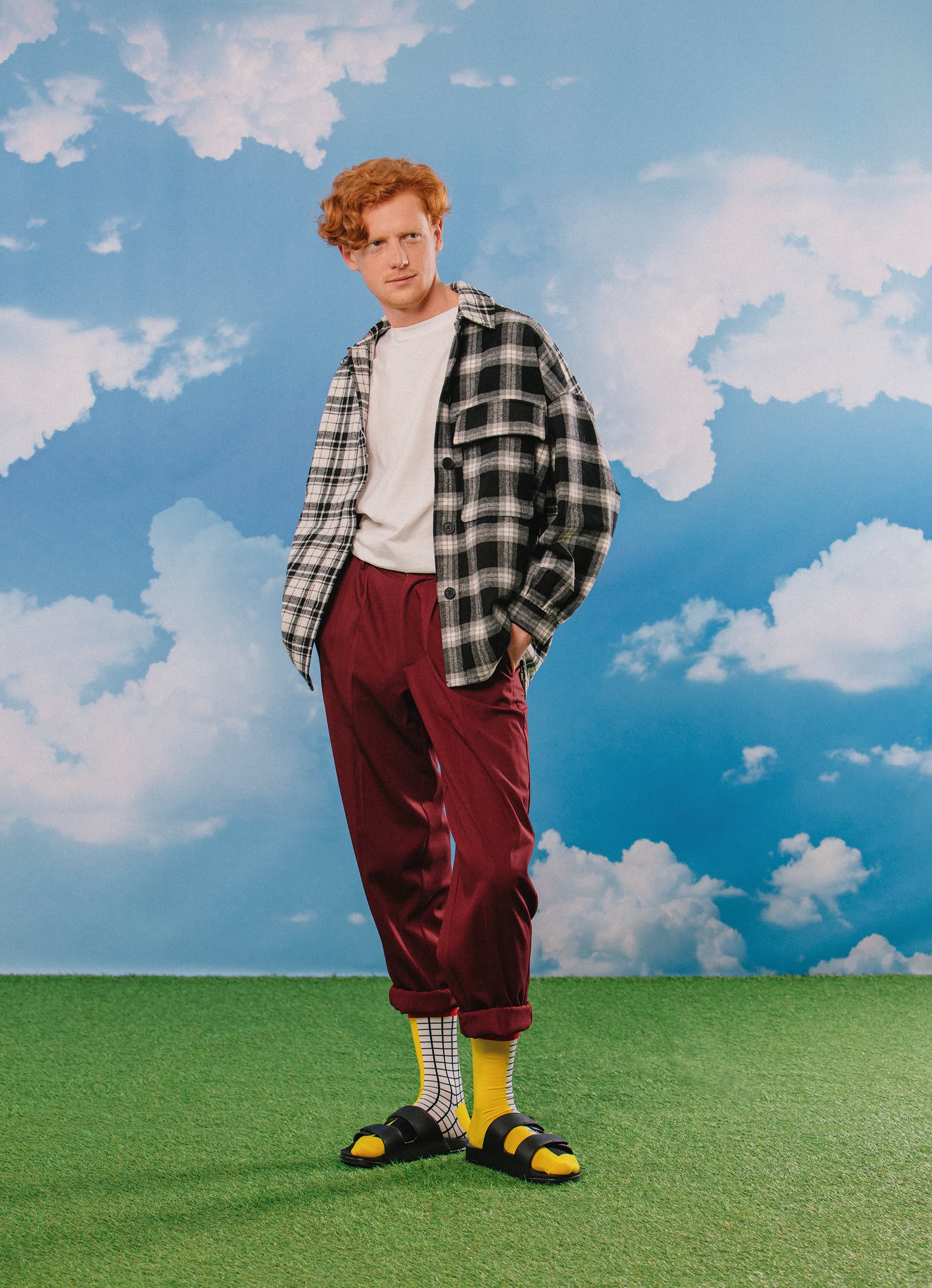 A curly red-haired person stands on green grass under a cloudy sky, wearing Eat My Socks French Fries Socks by Eat My Socks, a black-and-white plaid shirt over a white tee, maroon pants, black sandals, and quirky accessories.