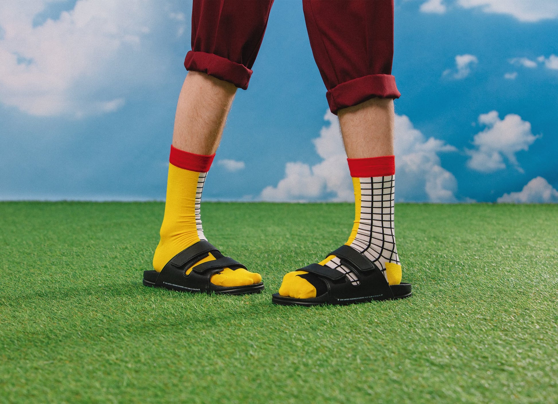 A person stands on artificial grass, wearing red rolled-up pants and Eat My Socks French Fries Socks by Eat My Socks—yellow with one plain and one black grid pattern—paired with black sandals, set against a blue sky with white clouds.