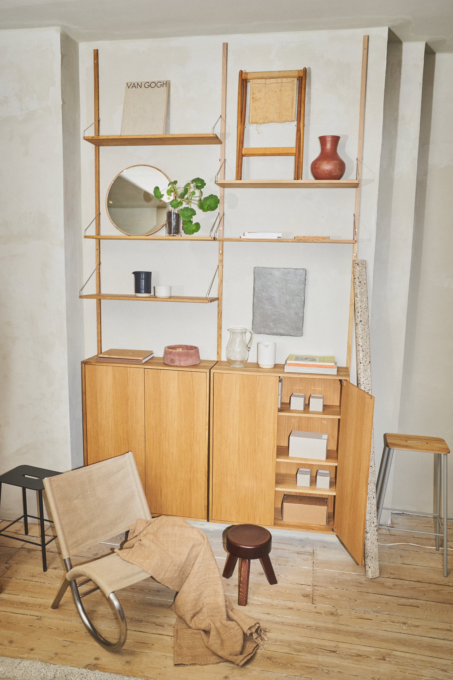 A solid oiled pine shelving unit holds books, a round mirror, plant, ceramic vase, and boxes against a light wall. The scene features the FRAMA AML Stool by FRAMA beside a low chair and draped fabric on the wooden floor for added versatility.