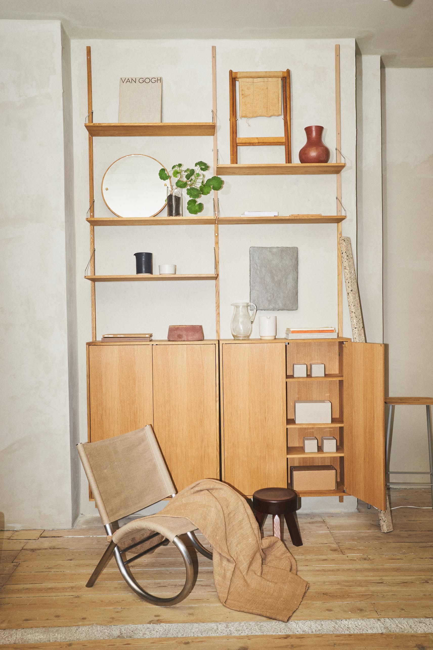 Minimalist interior with wooden shelves, ceramics, books, and a small plant. A modern lounge chair and the FRAMA AML Stool by FRAMA in solid oiled pine add versatility, all set on a warm wooden floor.