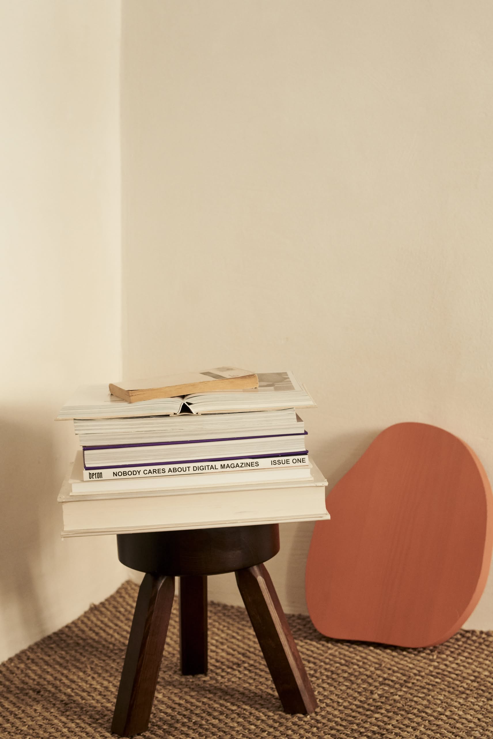 A compact FRAMA AML Stool in solid oiled pine holds books and magazines in a minimalist room with beige walls and a woven rug. Nearby, a flat, reddish-brown decorative piece rests against the wall.