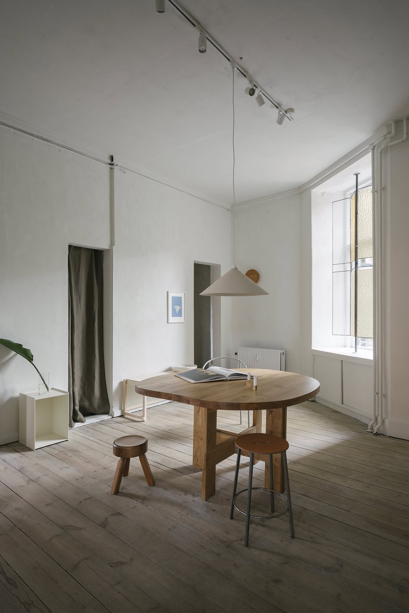 Minimalist room with a round wooden table, a solid oiled pine FRAMA AML Stool by FRAMA beside the bed, another stool, pendant lamp, plant in the corner, light wood floors, curtained doorway, and a tall window filling the space with natural light.