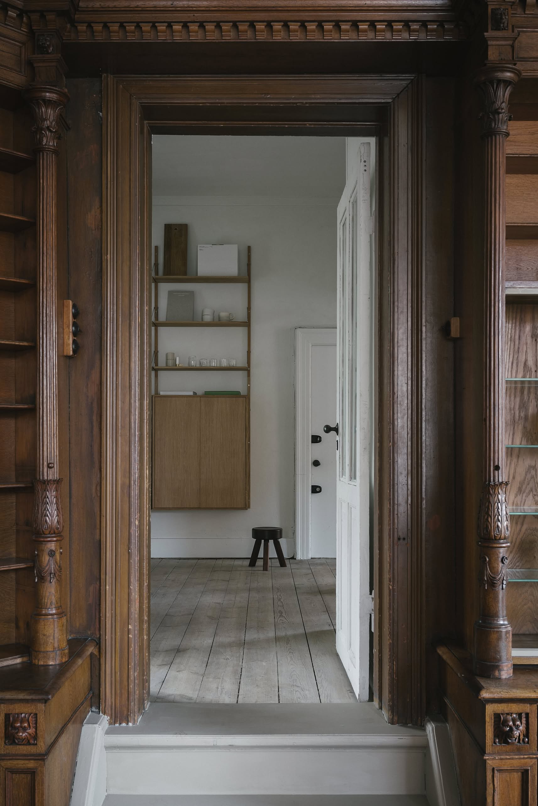 A view through a carved wooden doorway into a minimalist room with light wood floors, white wall, shelves, cabinet, and the FRAMA AML Stool by FRAMA—crafted from solid oiled pine—placed near a white door with black handles.