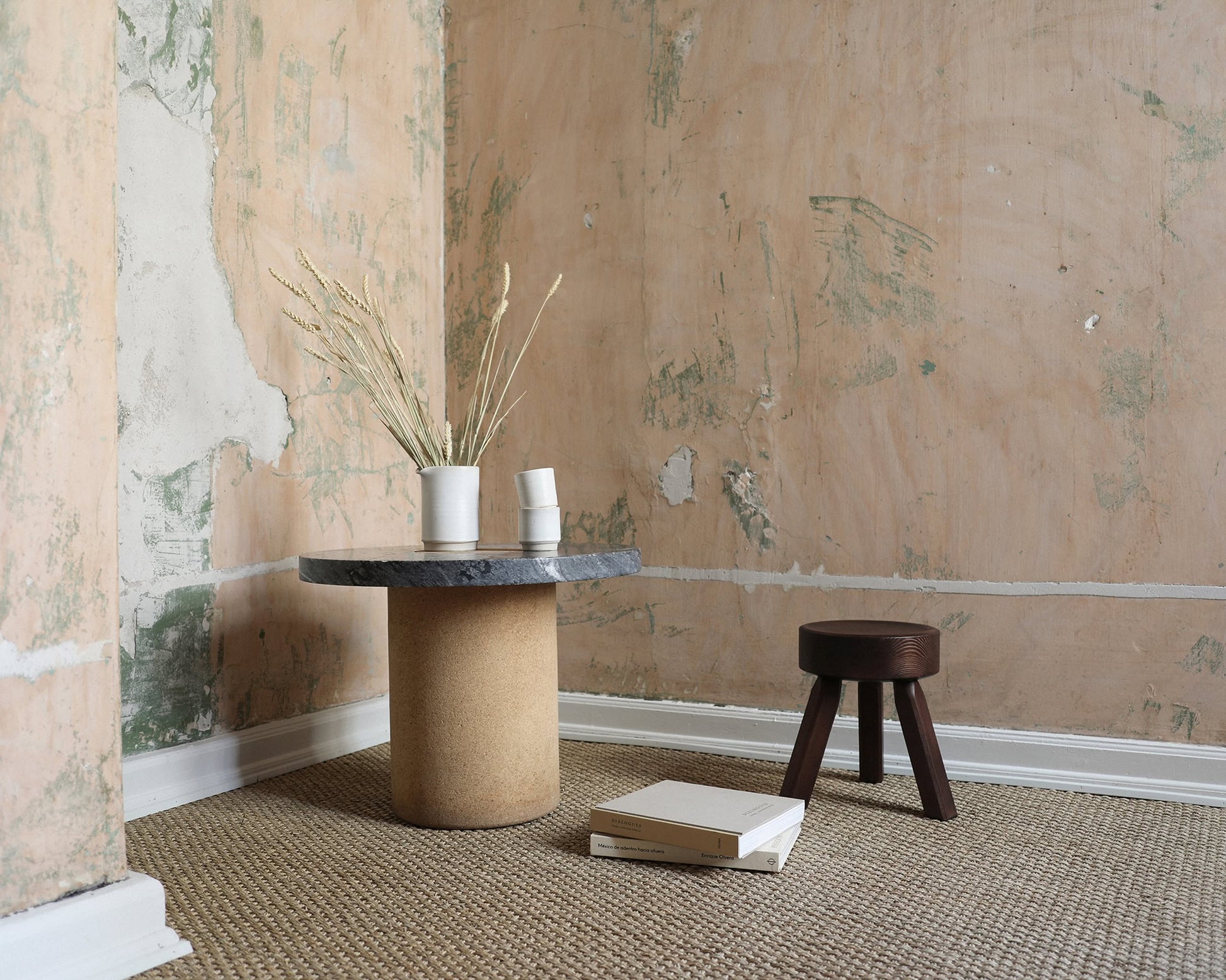 A minimalist room with peeling plaster walls features a round table with a vase of dried grasses and two cups, the FRAMA AML Stool by FRAMA crafted from solid oiled pine as a versatile bedside piece, and a closed book on a woven carpet.