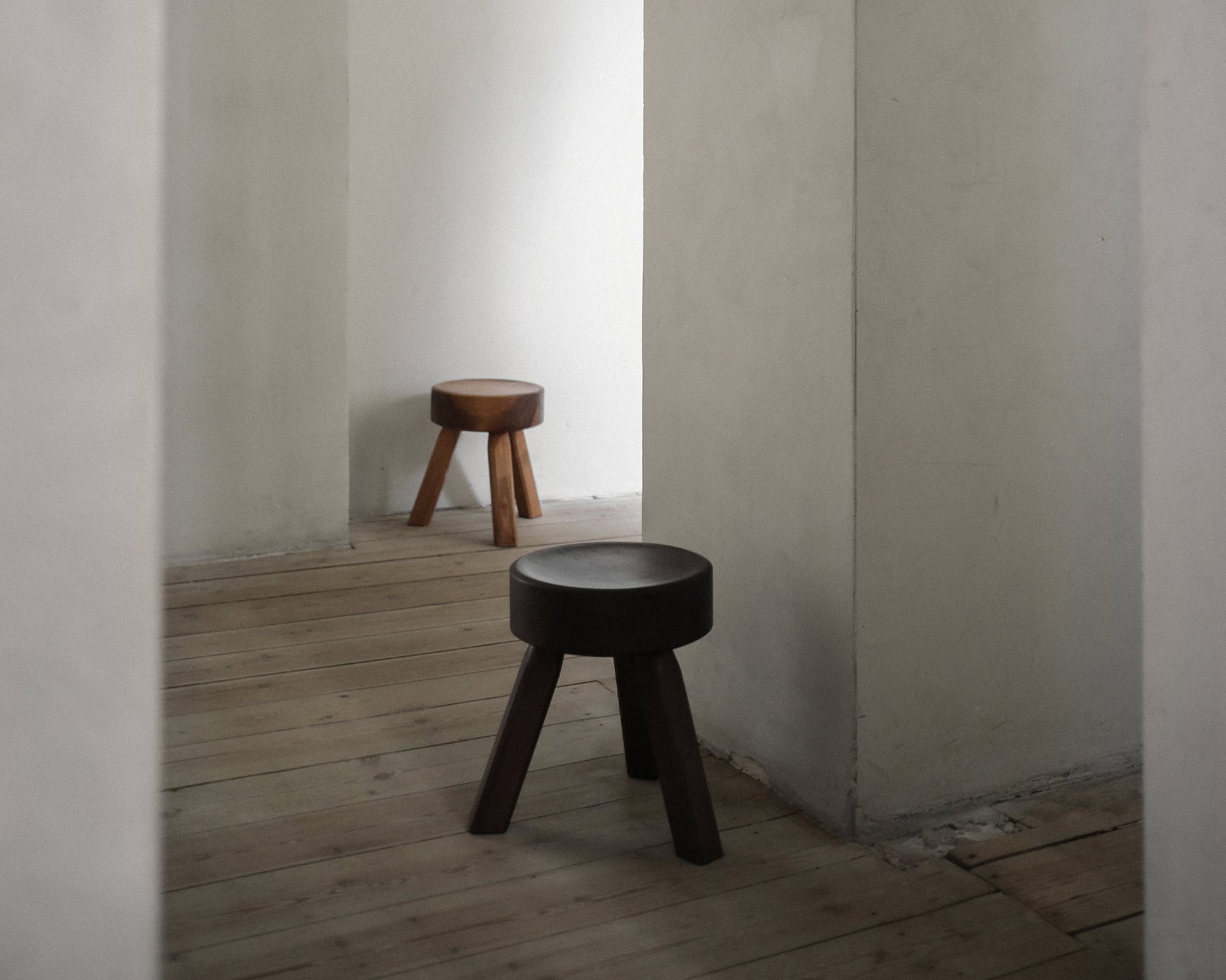 Two FRAMA AML Stools by FRAMA, one in dark and one in light solid oiled pine, stand in separate corners of a minimalist room. Their versatile design suits use as bedside stools or accent pieces, enhancing the space’s calm and simple vibe.