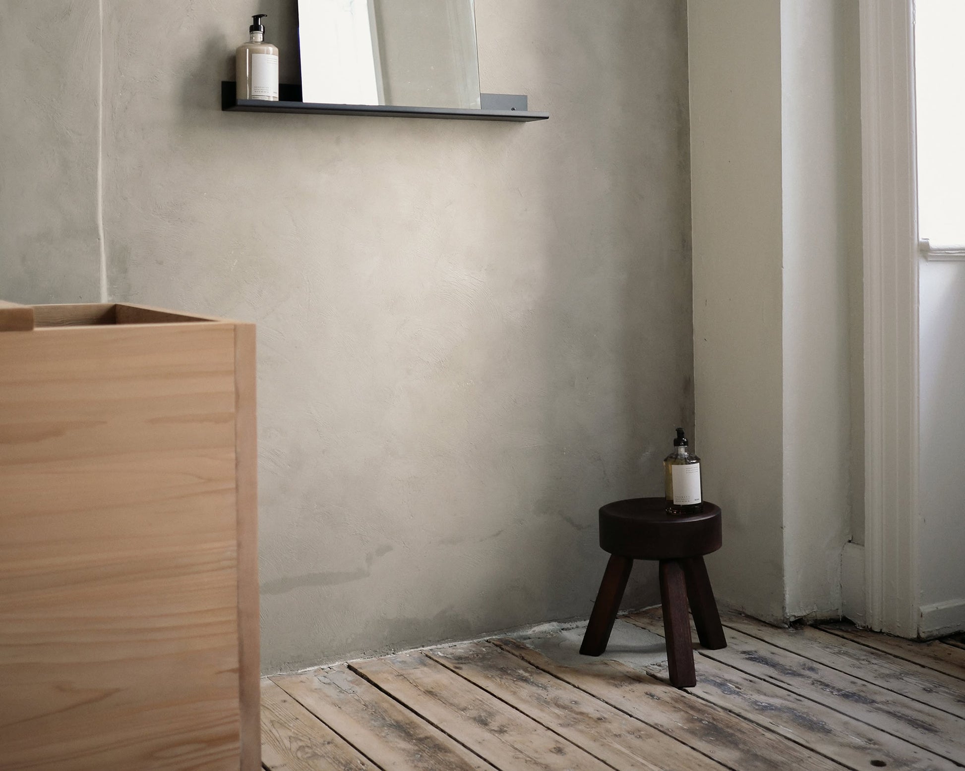 A minimalist bathroom with light wooden floors features a solid oiled pine cabinet and the FRAMA AML Stool by FRAMA as a side table holding a soap dispenser. A wall-mounted shelf with mirror enhances the versatile design, illuminated by window light on the right.