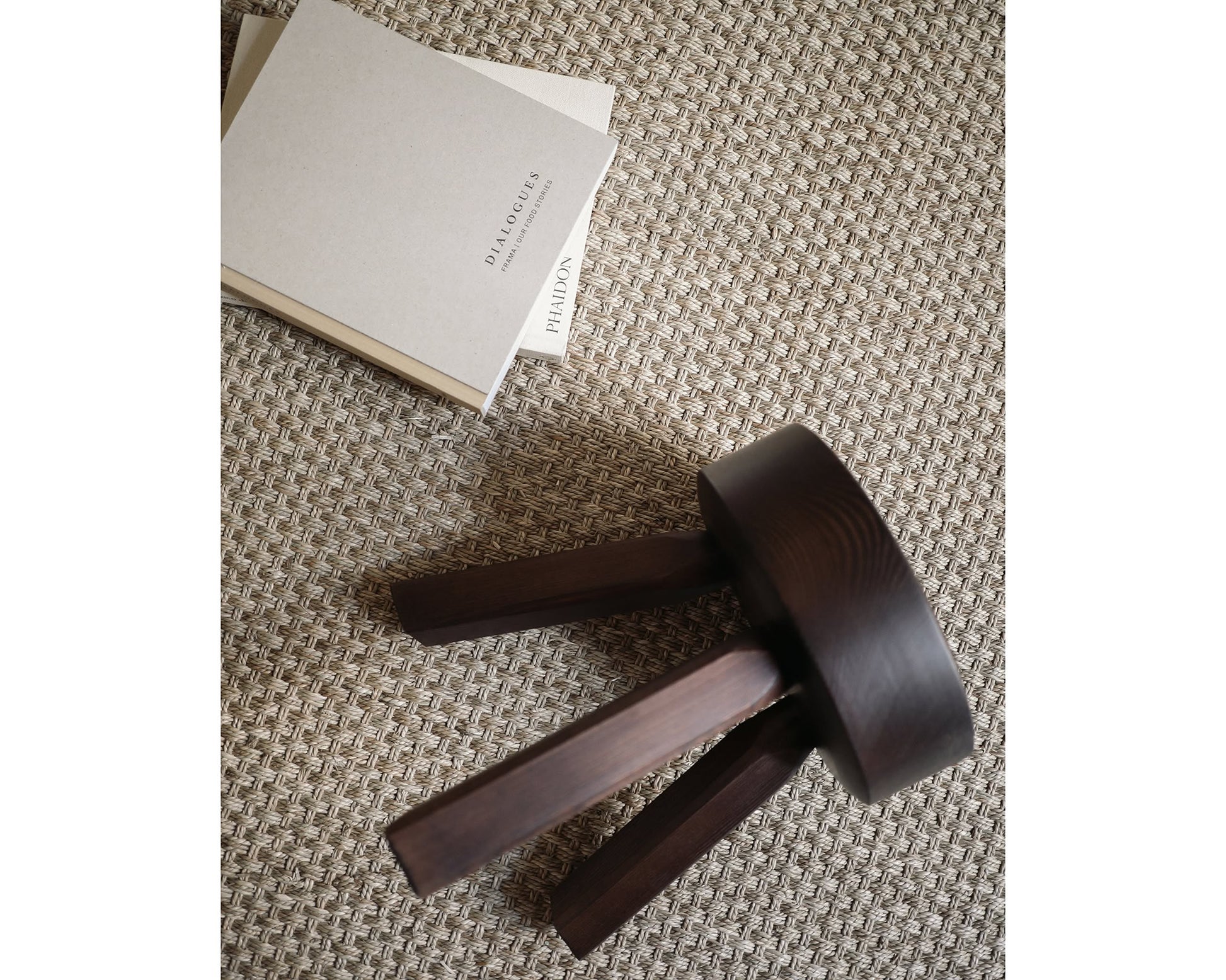 A compact, dark solid oiled pine FRAMA AML Stool by FRAMA rests on its side atop a textured beige carpet beside two closed, neutral-toned books, one titled DIALOGUES.