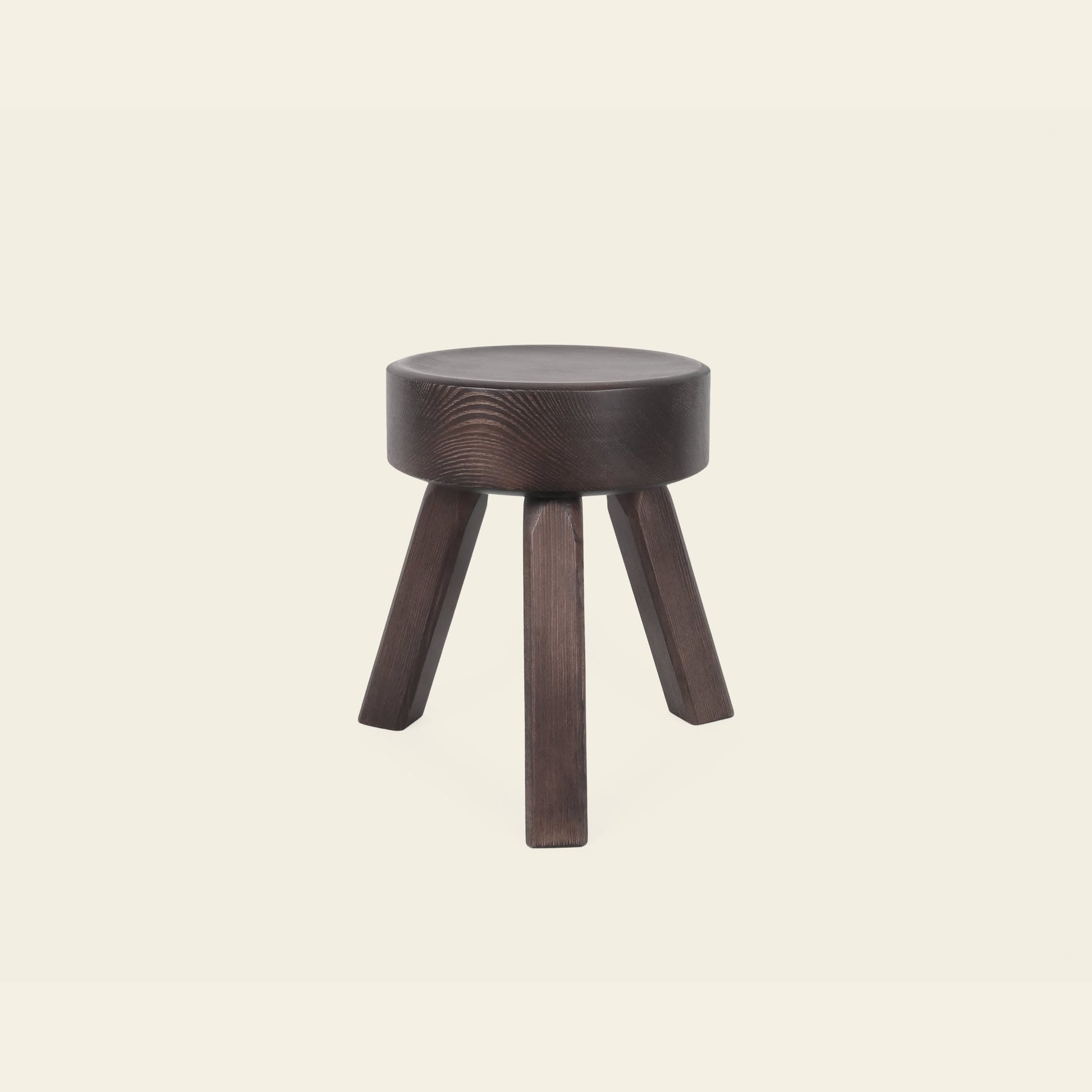 The FRAMA AML Stool by FRAMA features a small, round seat and three angled legs in solid oiled pine—ideal as a bedside stool. Its versatile, dark wood design stands out against a plain off-white background.