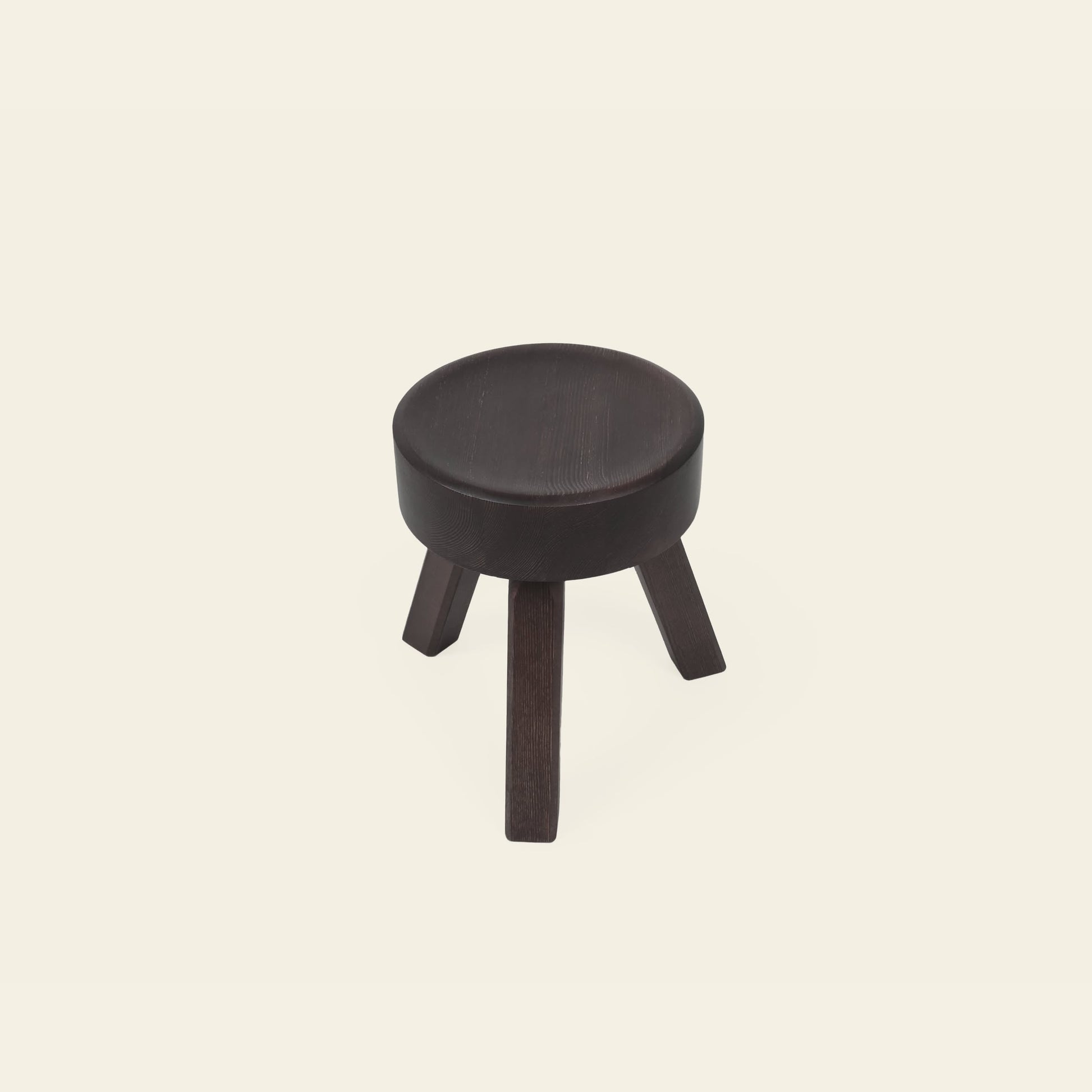 The FRAMA AML Stool by FRAMA is a small, round, dark wooden bedside stool with three angled legs, crafted from solid oiled pine and shown against a plain off-white background.