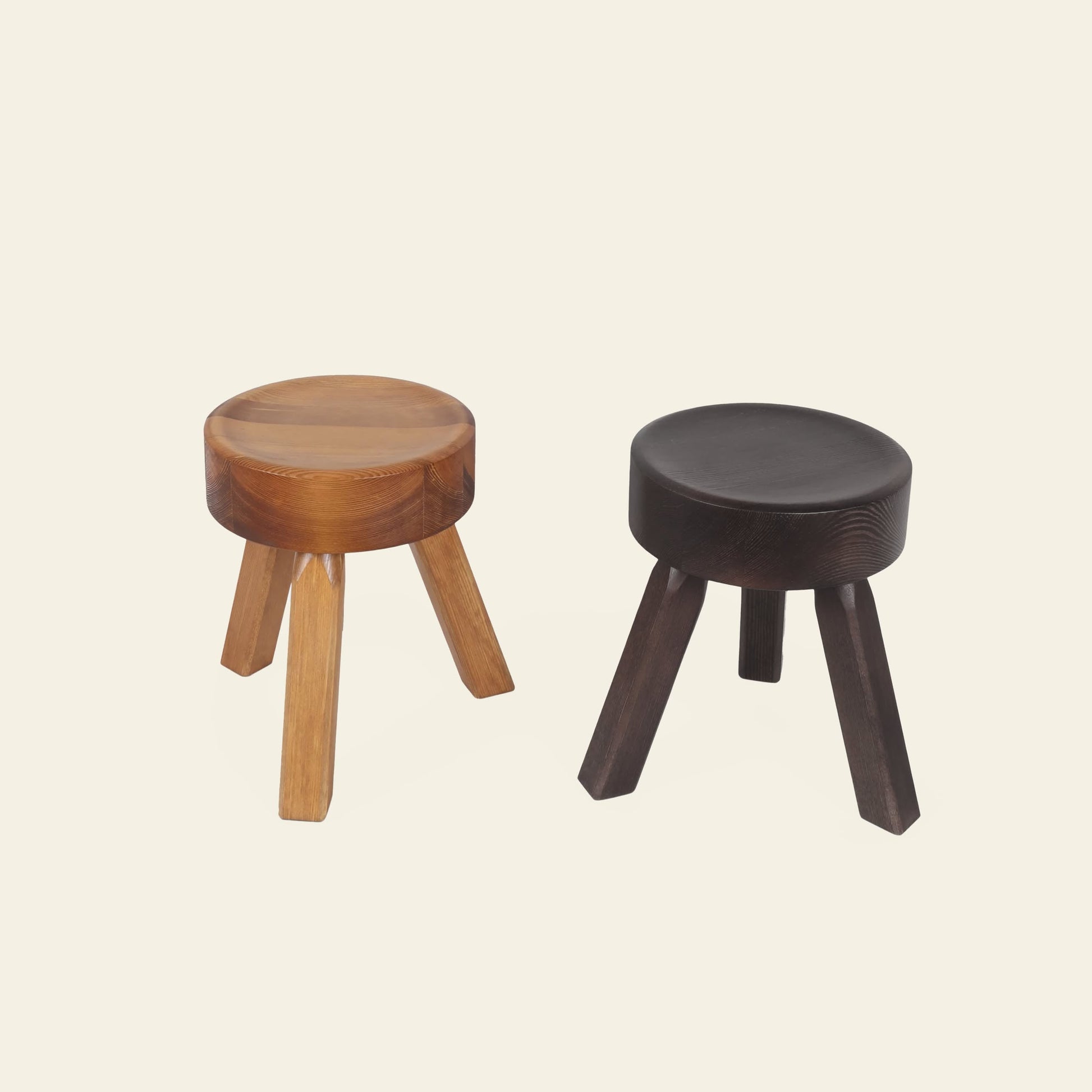 Two FRAMA AML Stools by FRAMA, each with a round seat and three legs in solid oiled pine—one in light natural wood, the other in dark brown/black—are shown on an off-white background, ideal for use as bedside stools.